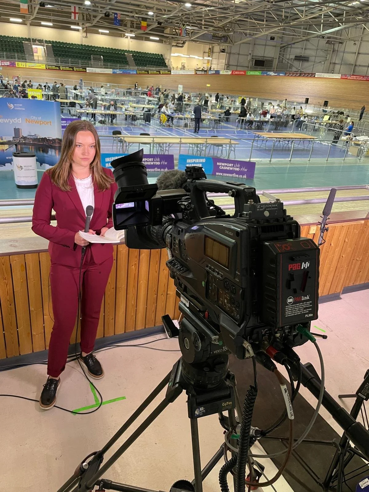 A news reporter stands with a microphone in her hand, wearing a maroon suit and black shoes, behind a camera on a tripod, in a sports hall where an event with tables and people is taking place in the background.