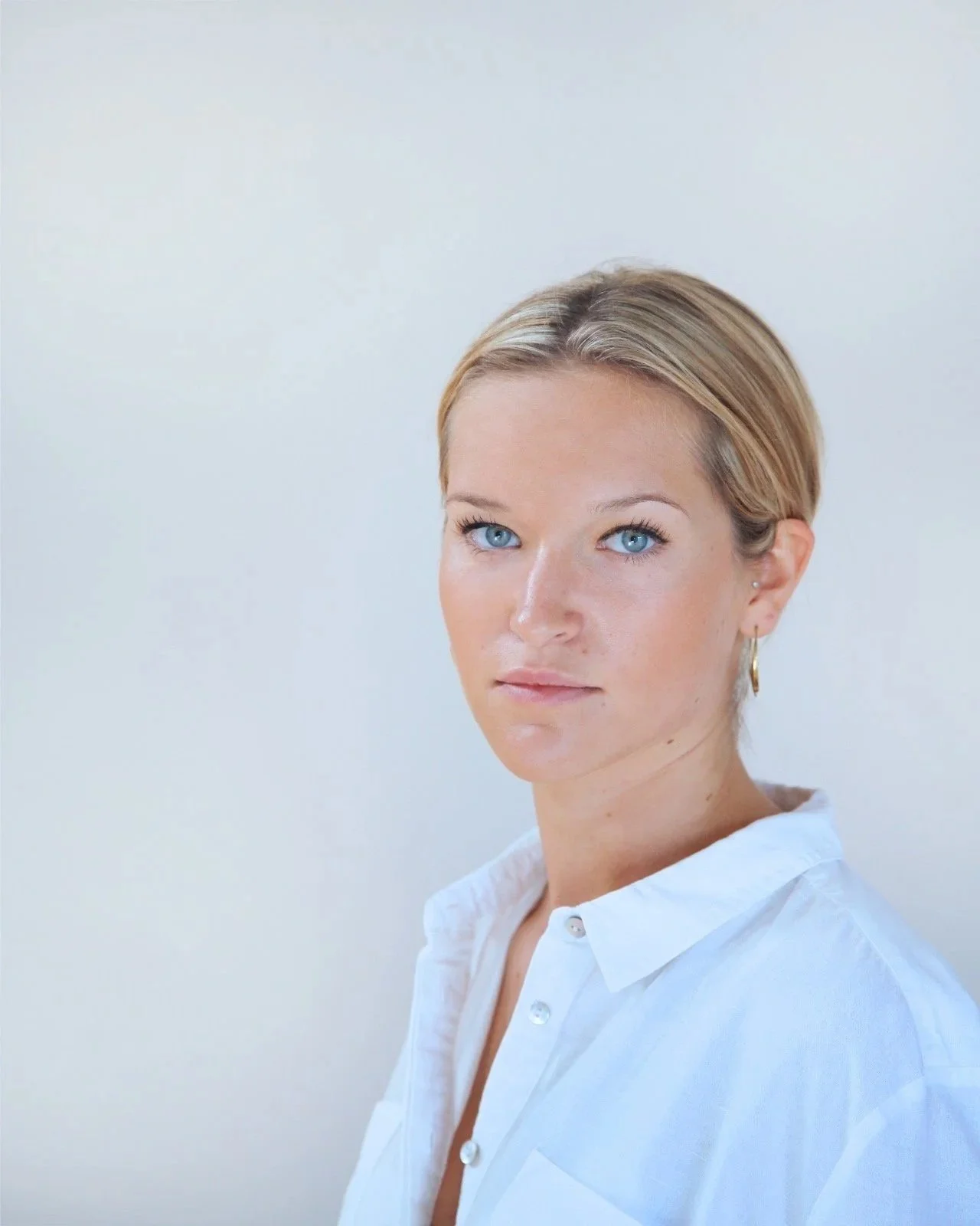 A woman, Alex Mears-Jennings, with blonde hair and blue eyes wearing a white button-up shirt, standing against a plain white background.