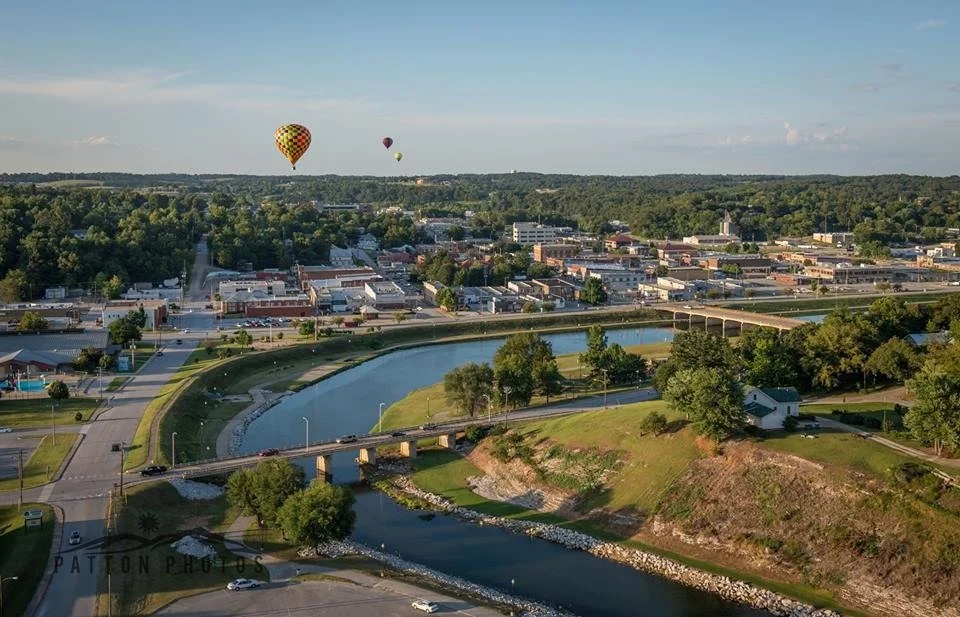 Aerial view of a town with a winding river, a bridge, trees, and several hot air balloons floating in the sky.