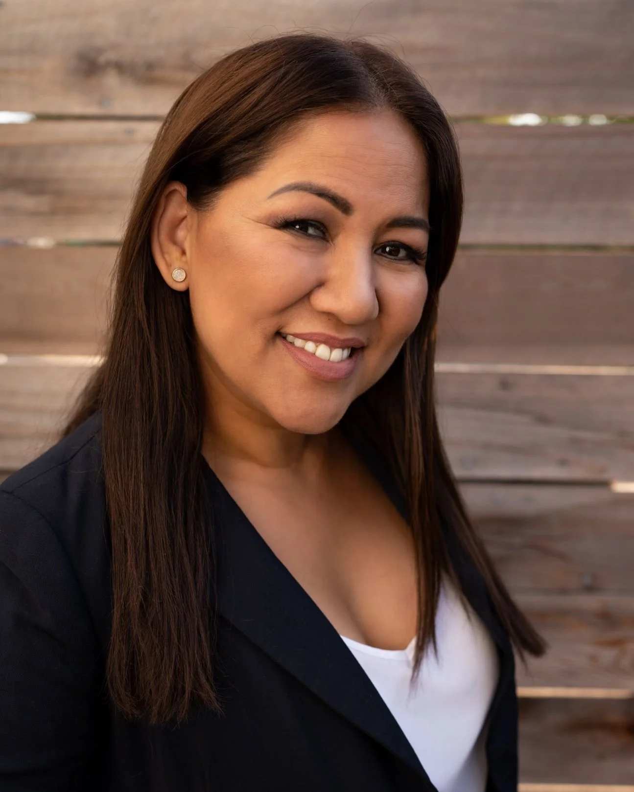 A woman smiling, with long brown hair, wearing earrings, a white shirt, and a dark blazer, standing outdoors in front of a wooden fence.