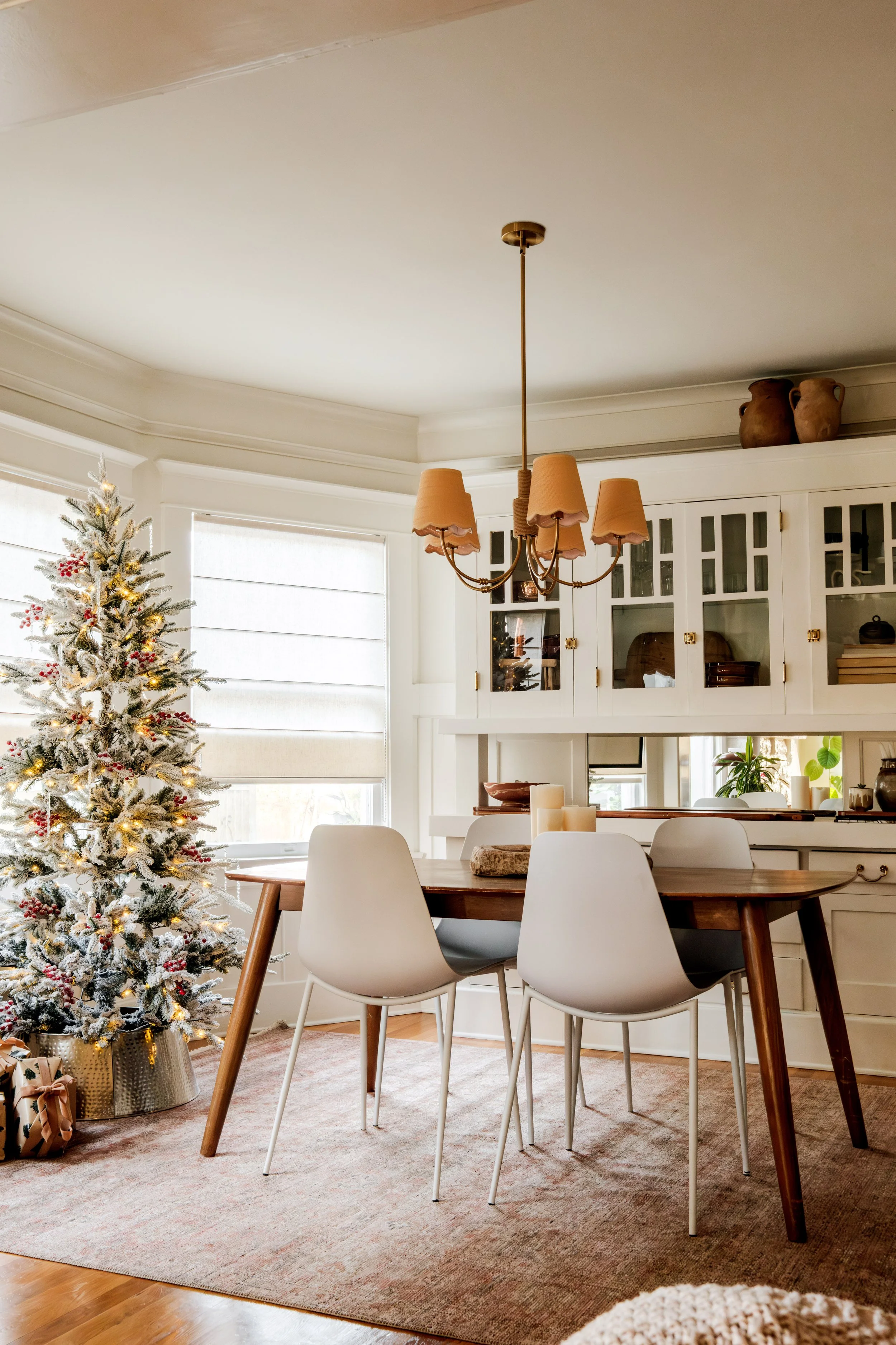 A dining room with a Christmas tree, a wooden dining table, white chairs, a beige rug, and a chandelier with tan lampshades.