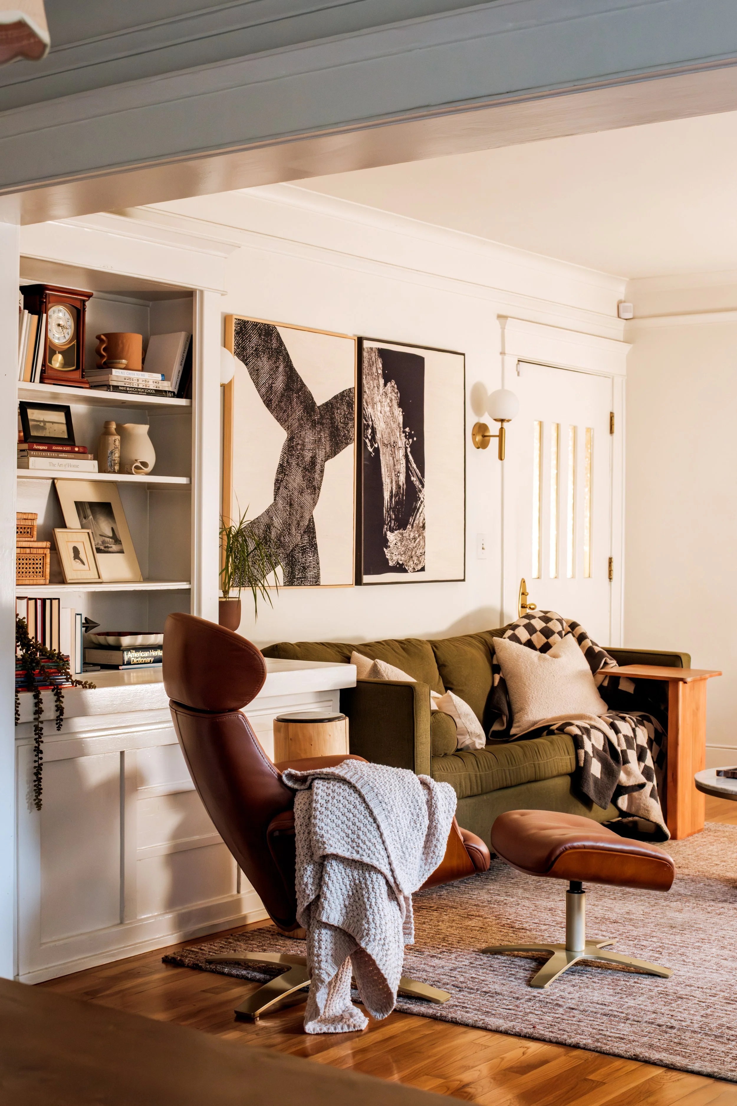 Living room with a brown leather lounge chair and matching ottoman, a green couch with cushions and a blanket, a built-in bookshelf with books and decor, black-and-white artwork, a wooden side table, and hardwood flooring.