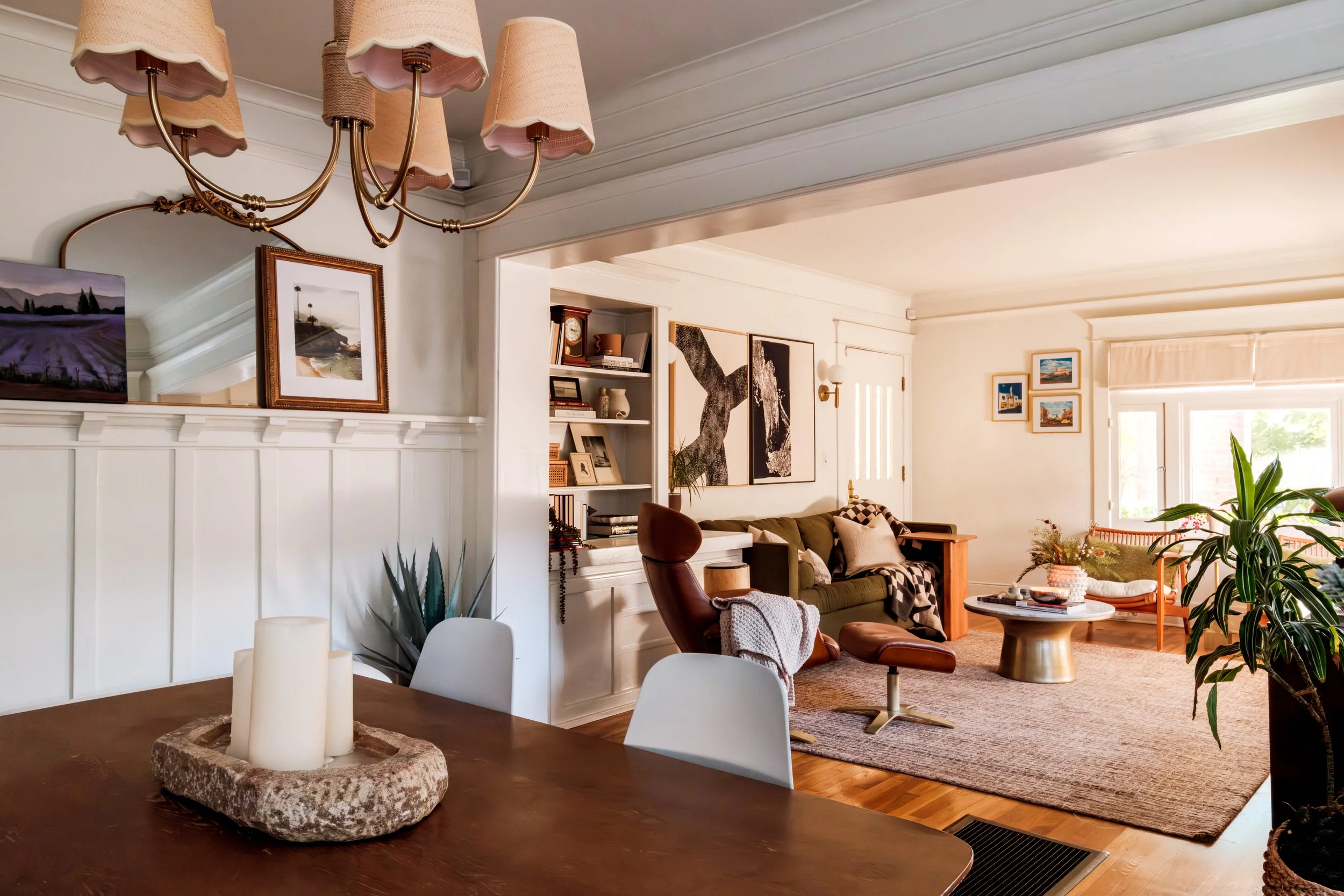Interior of a cozy living room with a dining area in the foreground, featuring a wooden table with candles. The living room has a green sofa, a brown leather chair, and a wooden bench, with artwork hanging on the white walls. Large windows let in nat