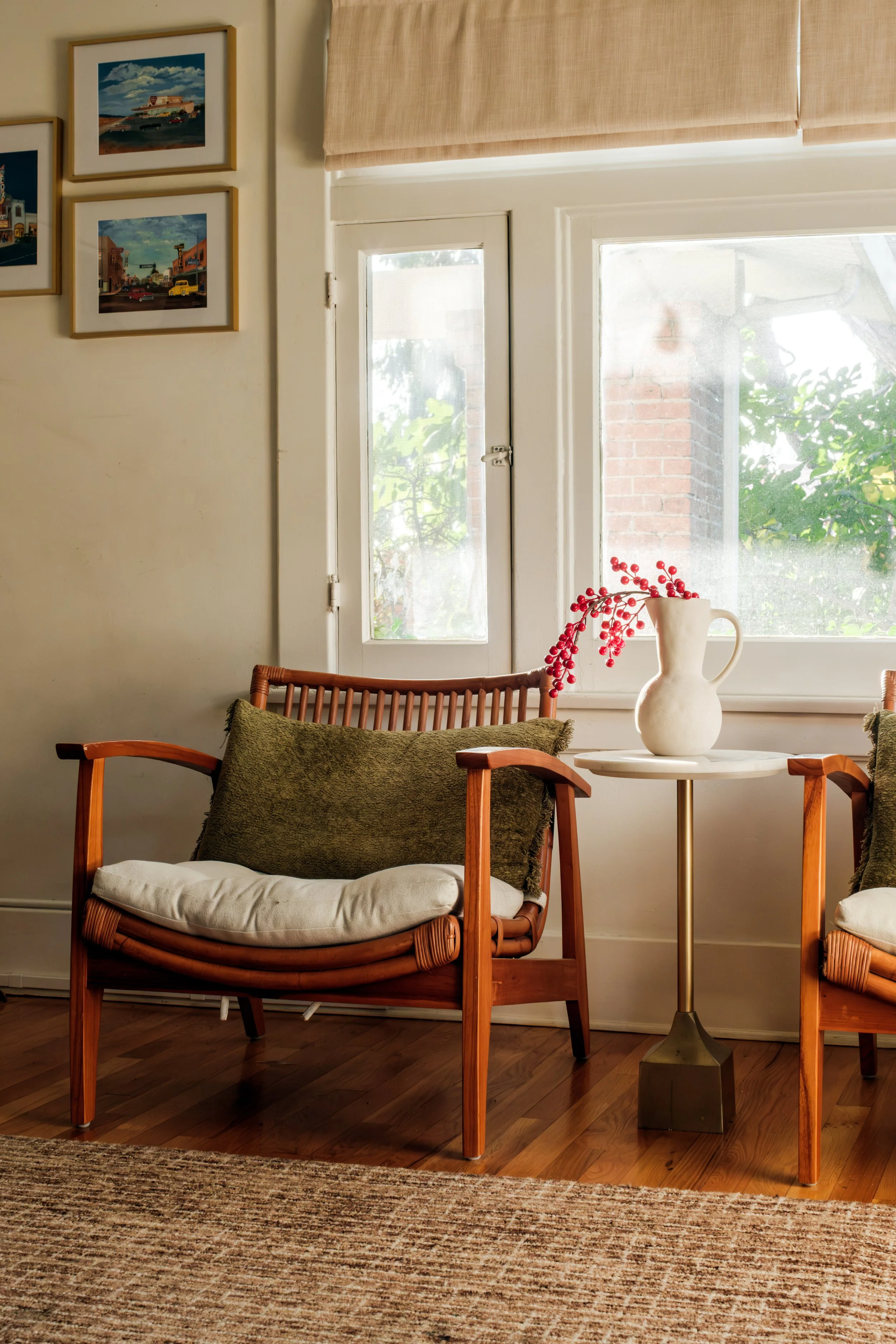Living room with two wooden chairs featuring cushions, a white side table with a white vase containing red berries, and framed artwork on the wall. Bright window with beige blinds revealing greenery outside.