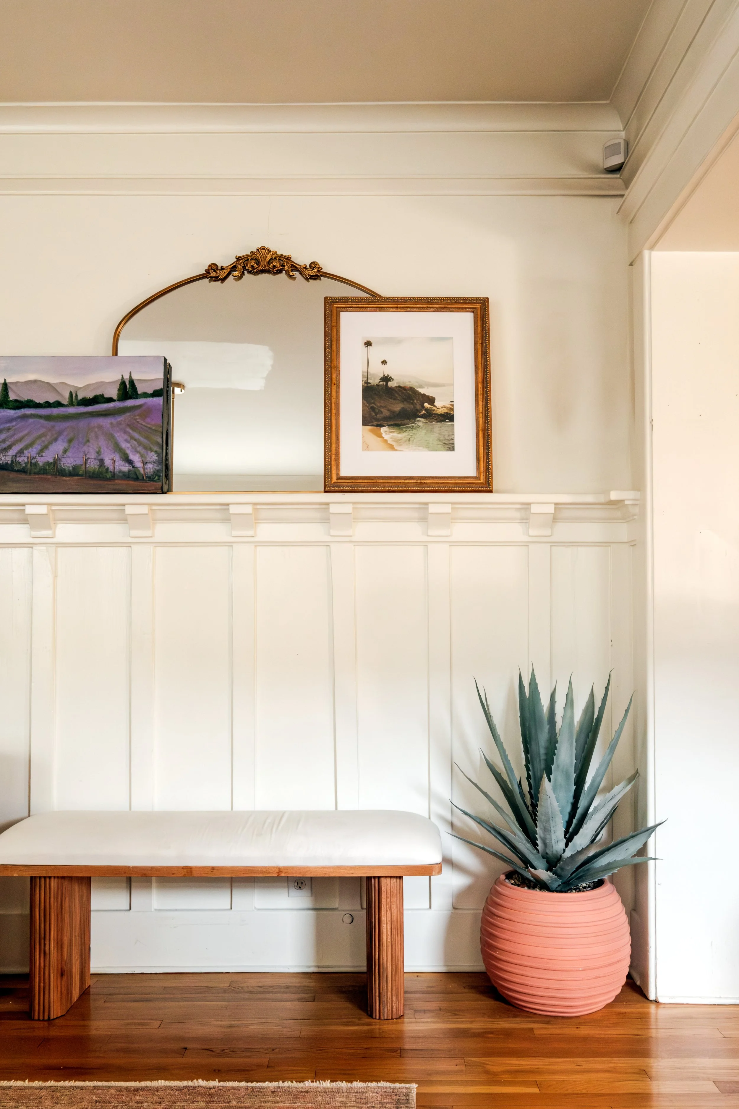 A white bench with wooden legs in front of a white wall, with a large agave plant in a pink pot on the right, and artworks including a painting of lavender fields and a framed sunset photograph above.