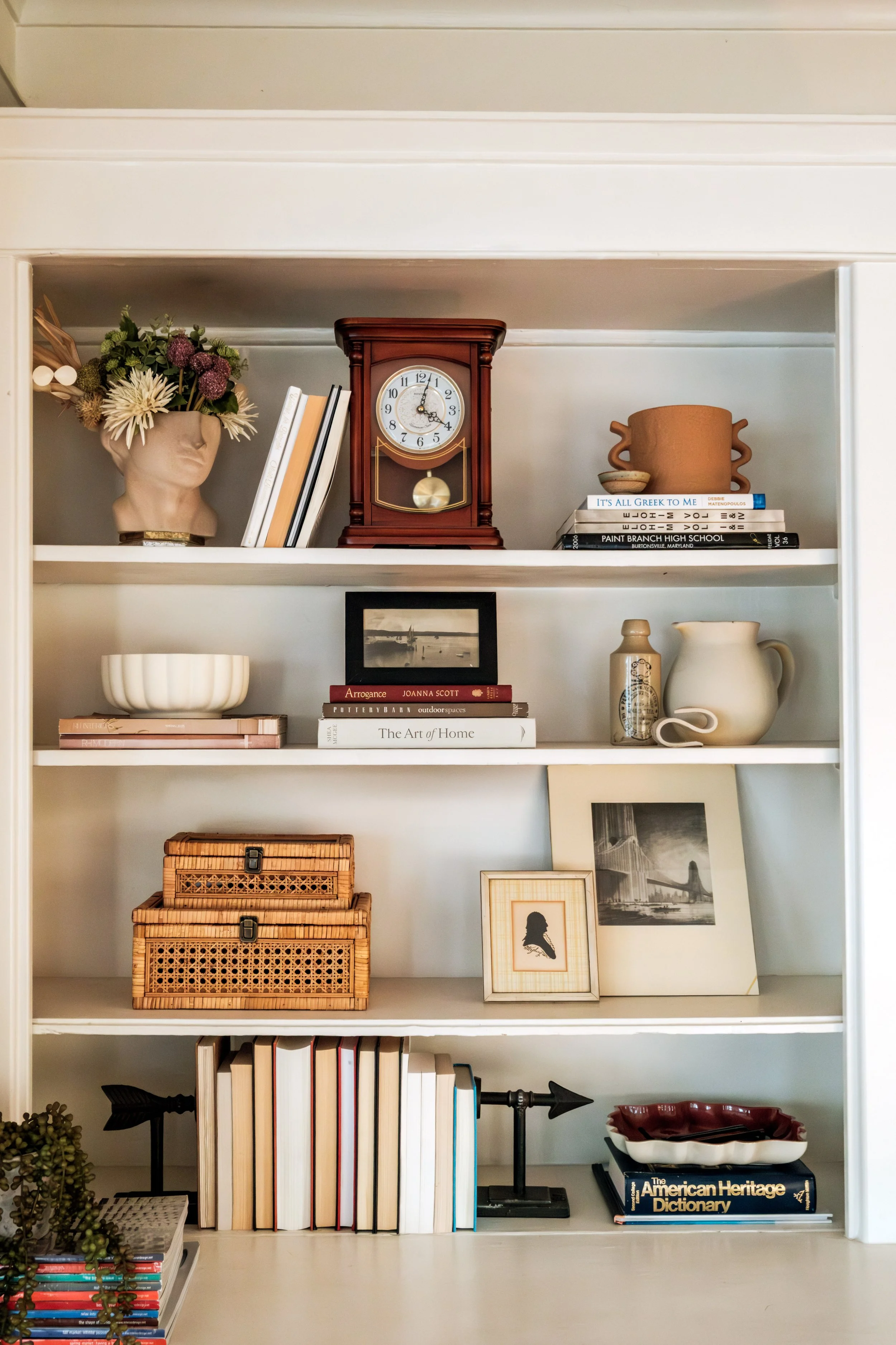 Shelves with books, decorative items, and framed photos, including a flower head sculpture, a red clock, ceramic pitchers, and woven storage boxes.