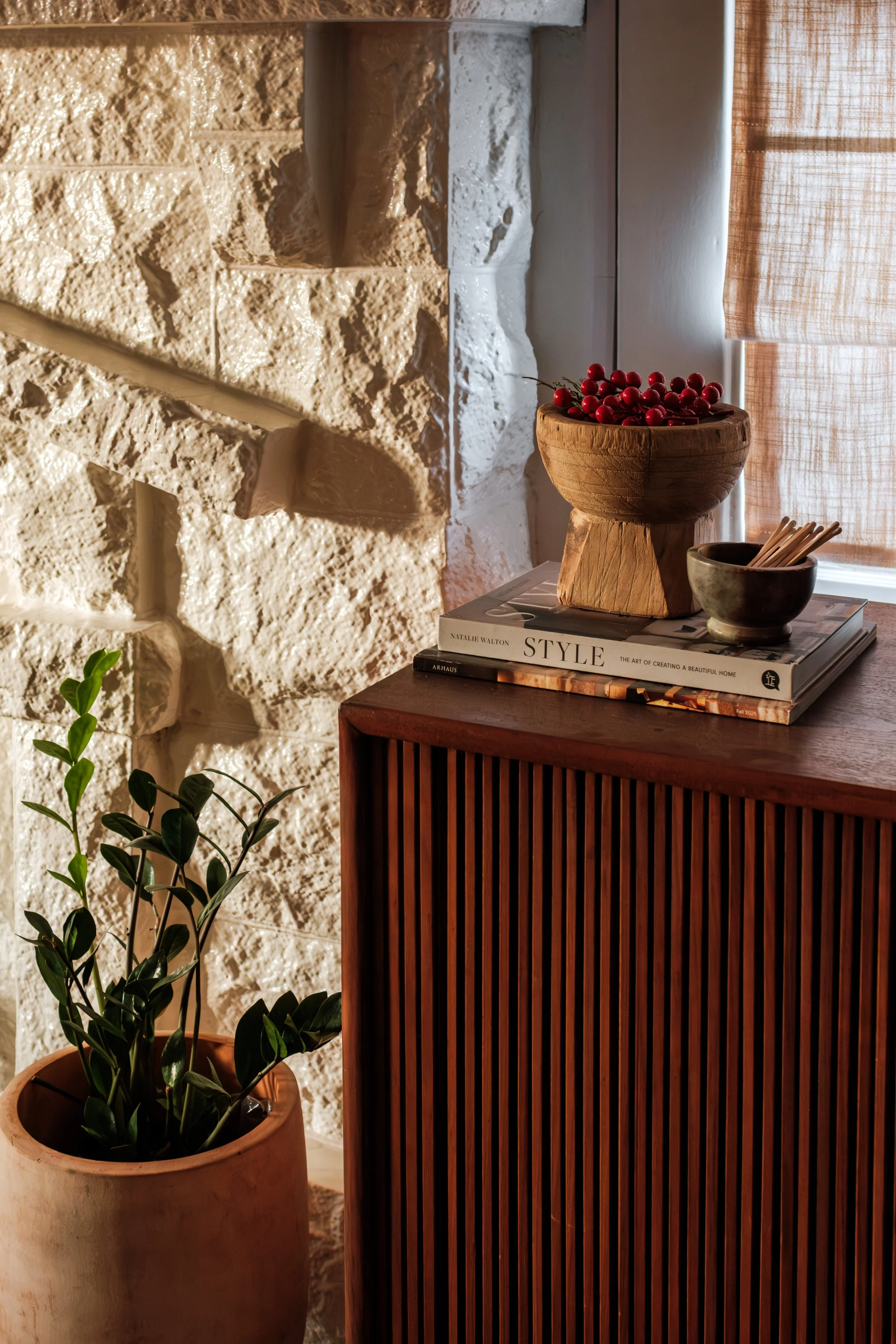 Decorative interior scene with a wooden sideboard, a potted plant, and a stone wall, featuring a stack of books topped with a bowl of cherries and a small bowl with utensils near a window with light-colored curtains.