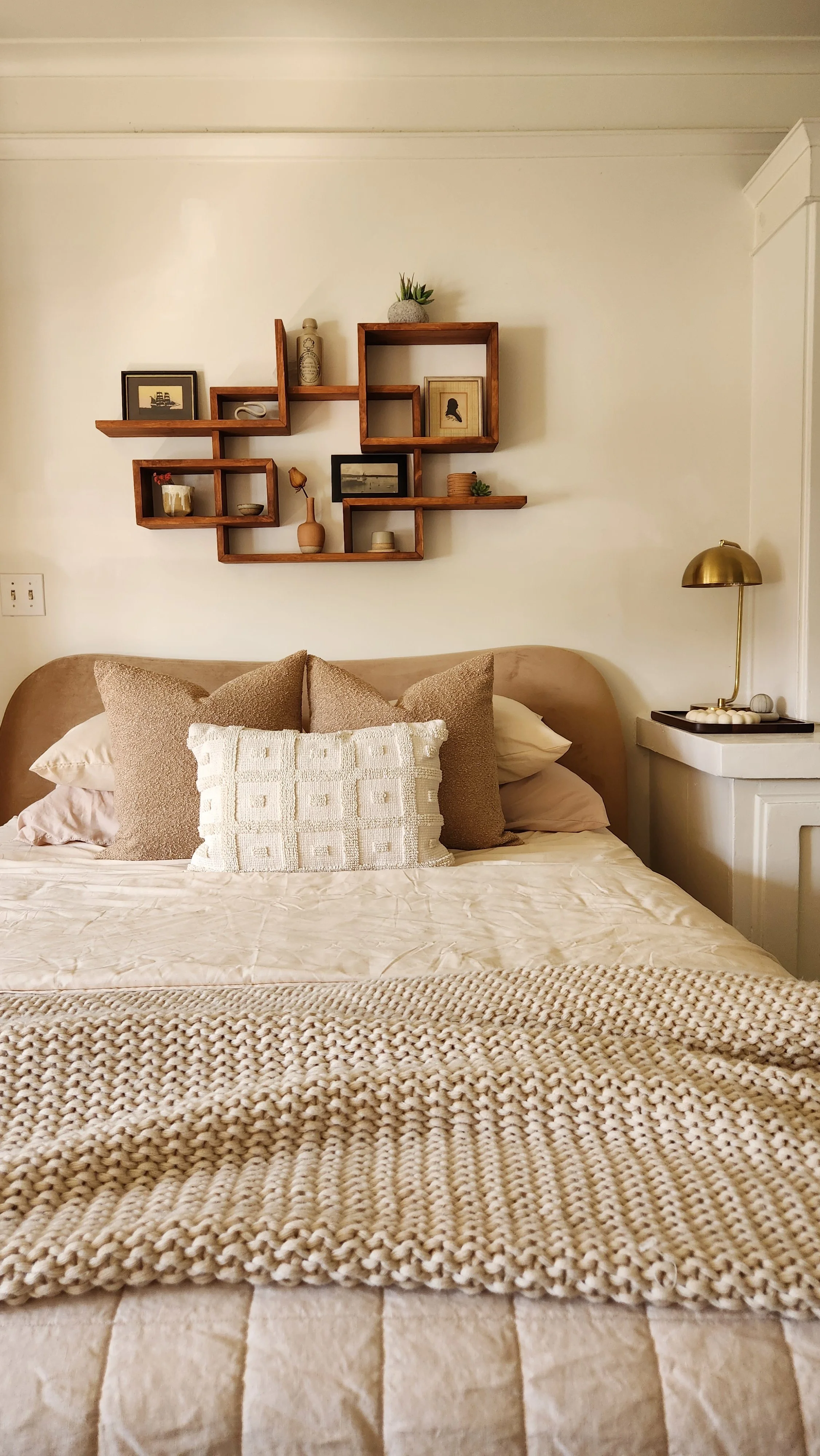 A cozy bedroom featuring a bed with multiple beige pillows and a chunky knit throw blanket. Above the bed, there is a wooden wall shelf with various decorative items, and a white nightstand with a gold lamp on the side.