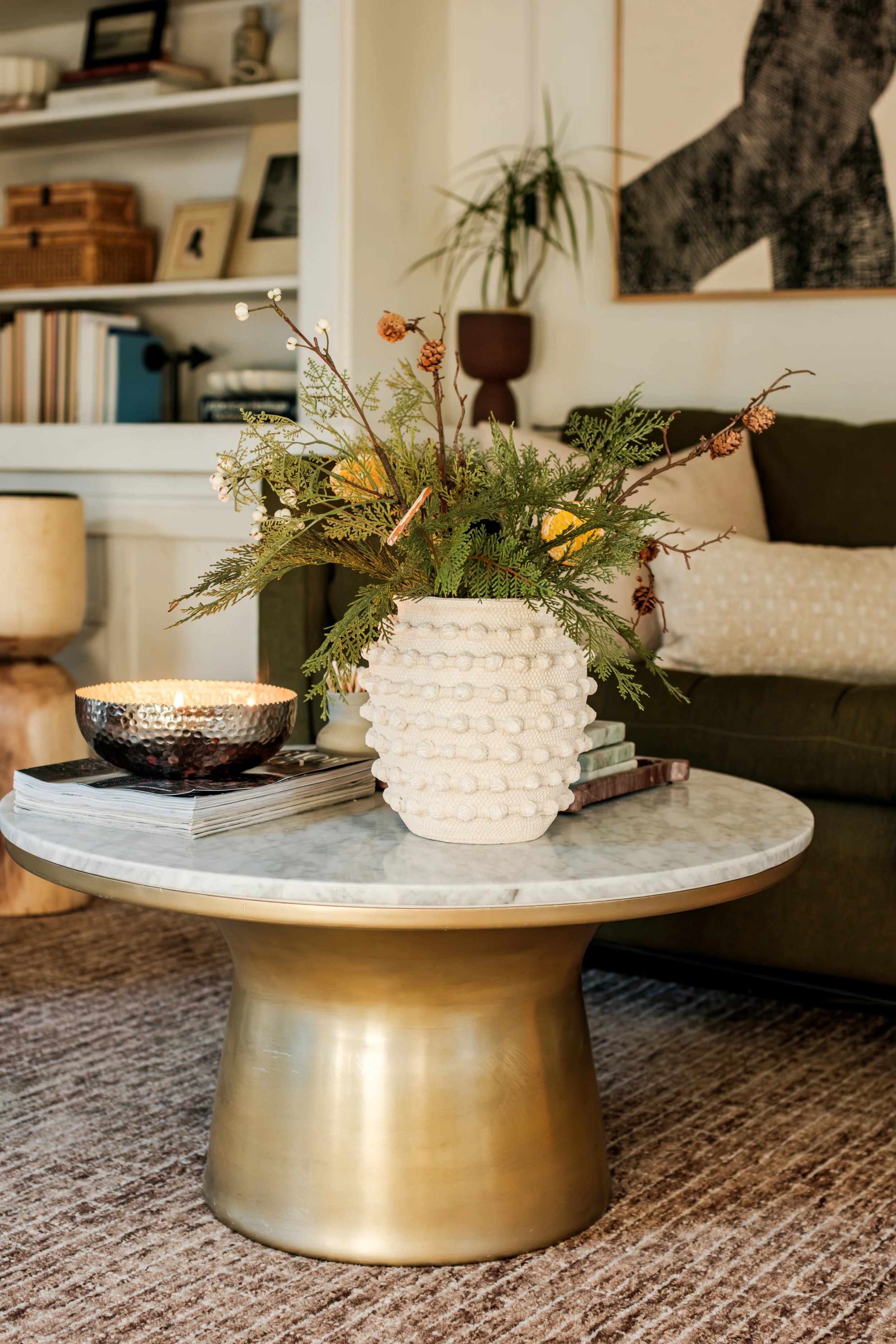A living room with a marble-top coffee table featuring a textured white vase with green foliage and pinecones, a black hammered metal bowl with a candle, and a few stacked books. A beige carpet and a dark sofa with pillows are visible in the backgrou