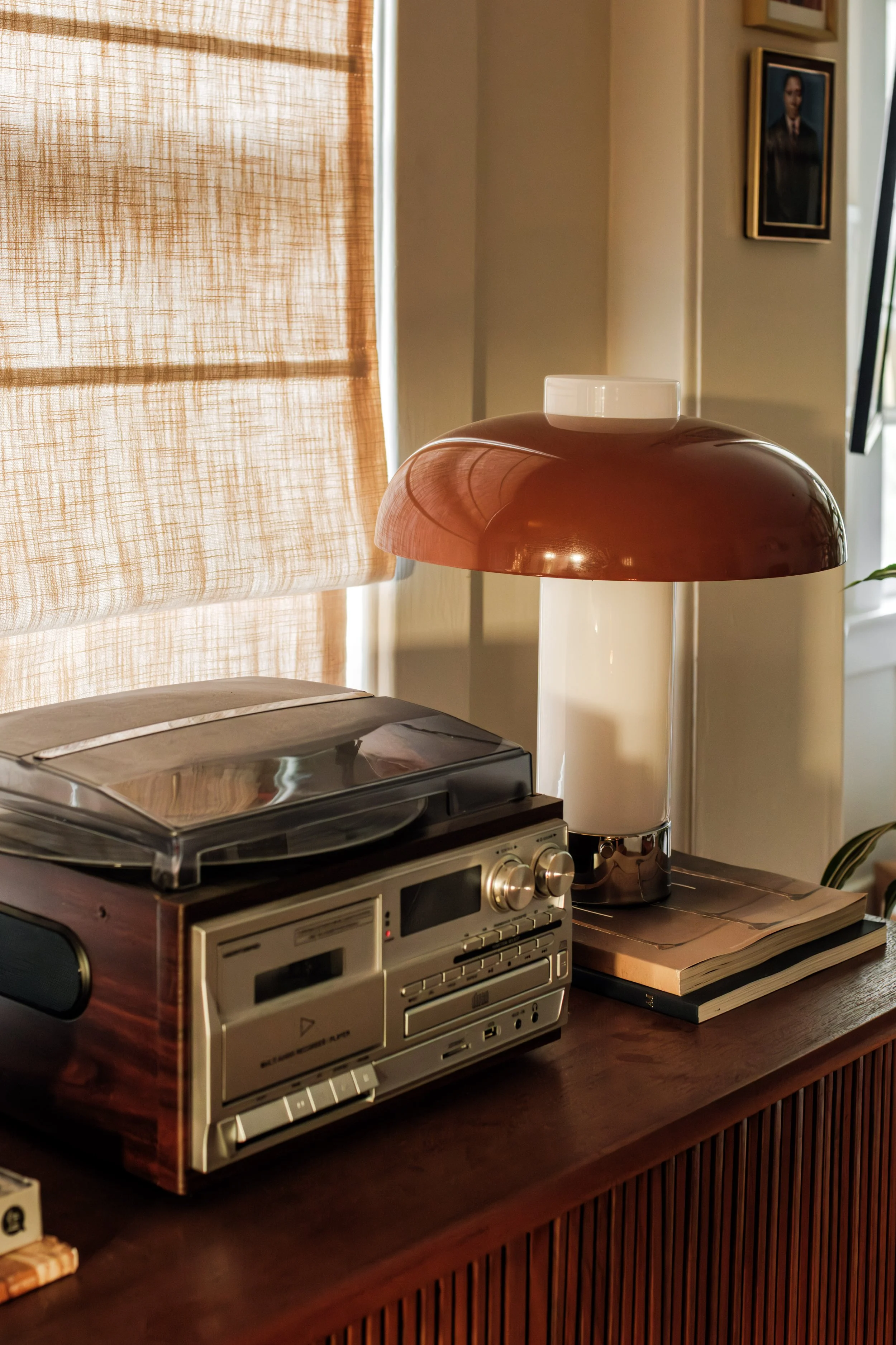 A vintage stereo with a turntable sits on a wooden cabinet next to a large white and brown lampshade, with framed photos hanging on the wall behind.