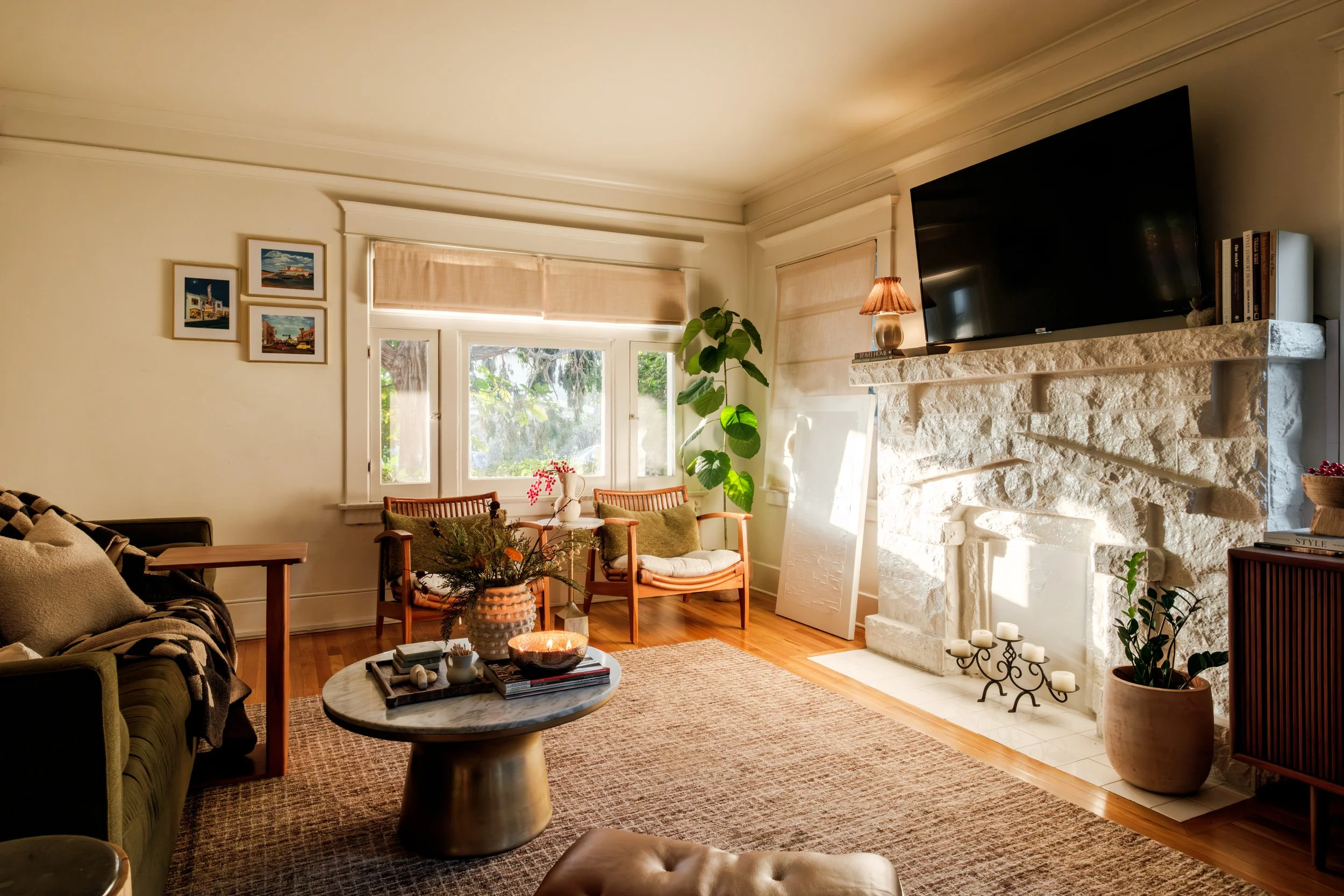 Bright living room with white stone fireplace, a large TV above, potted plants, wooden furniture, and natural light from windows.