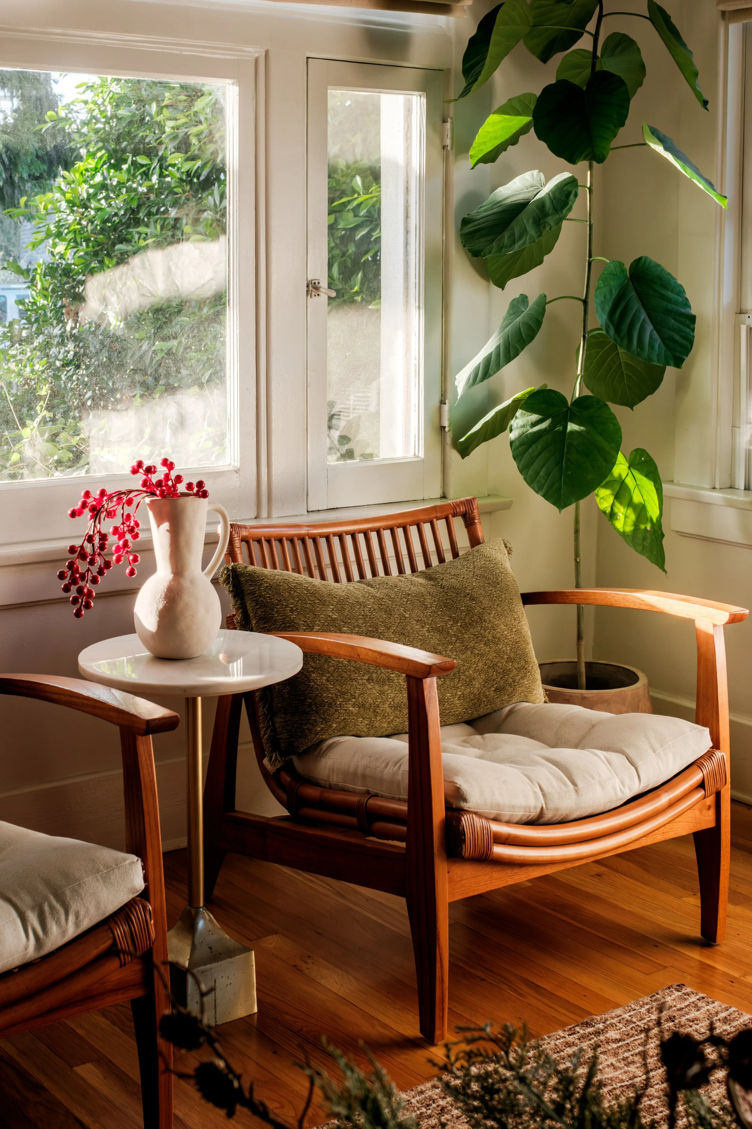 Sunlit interior corner with a wooden chair featuring a cushion and a pillow, a small white side table with a white ceramic vase holding red berries, large green leafy plant, window with sunlight streaming in, wooden floor, and part of a rug.