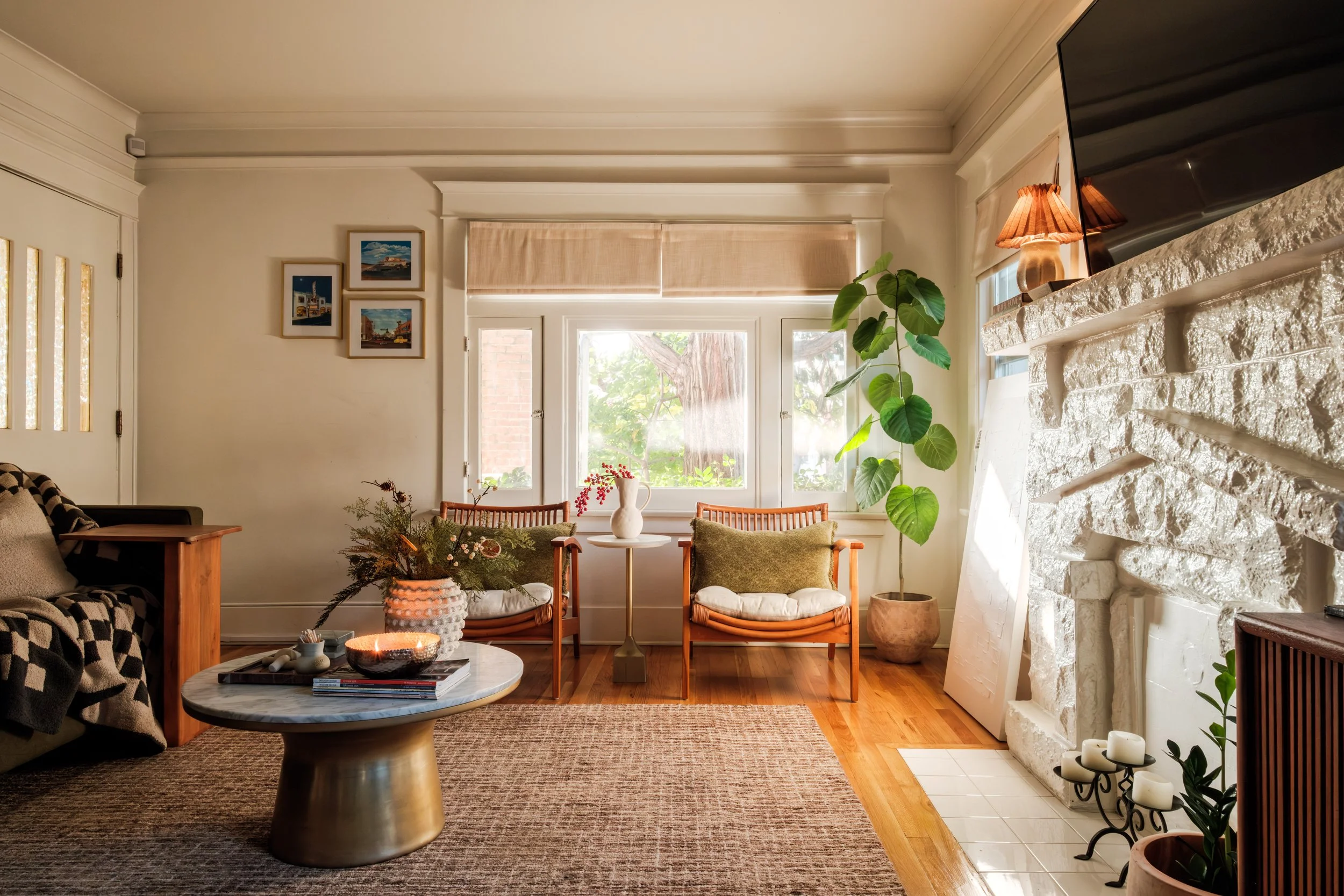 Living room with a fireplace, a large window, a beige wall, framed artwork, a black and wooden sofa, a round marble coffee table, two wooden armchairs with pillows, a tall potted plant, and decorative candles.
