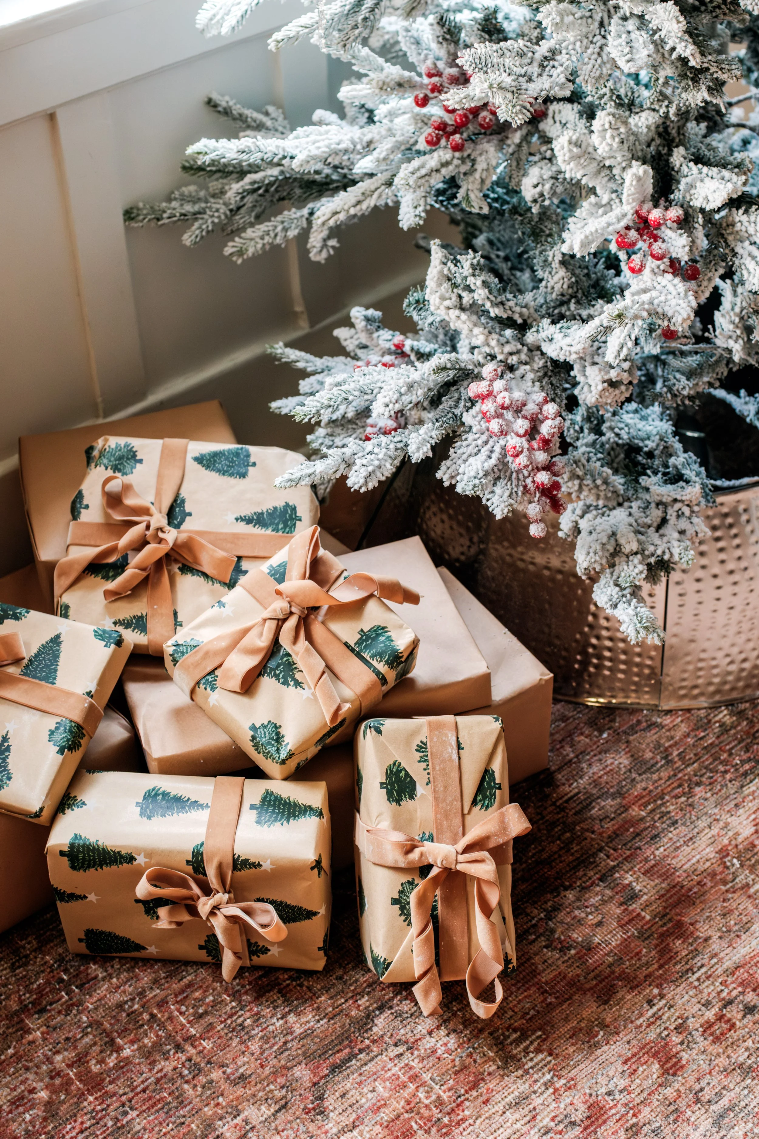Christmas presents wrapped in holiday paper with bows under a decorated snow-frosted Christmas tree.