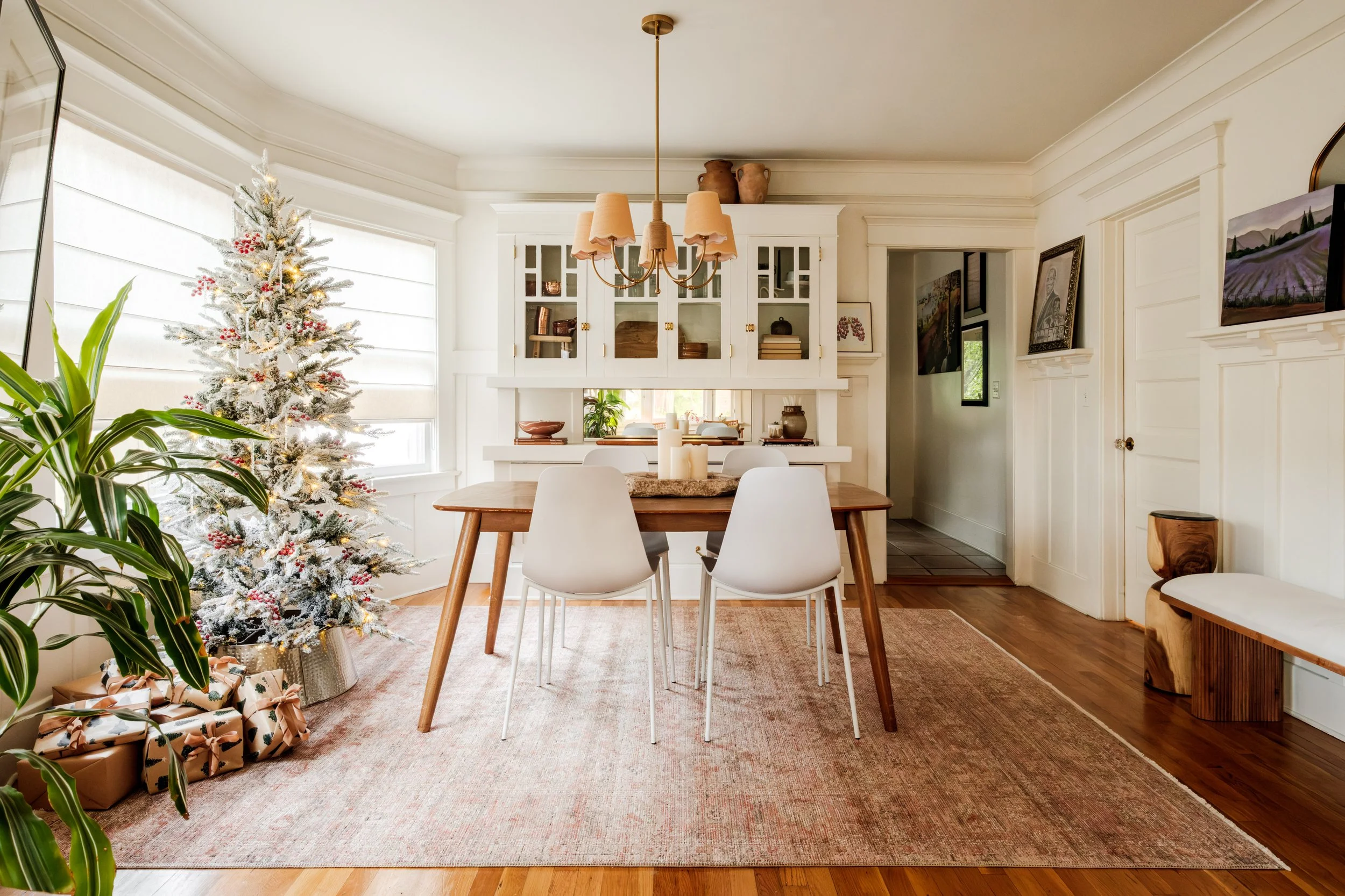 A Christmas tree decorated with white and red ornaments and surrounded by wrapped gifts, placed in a bright dining room with white walls, a wooden table with white chairs, a pinkish rug, and a built-in cabinet in the background.