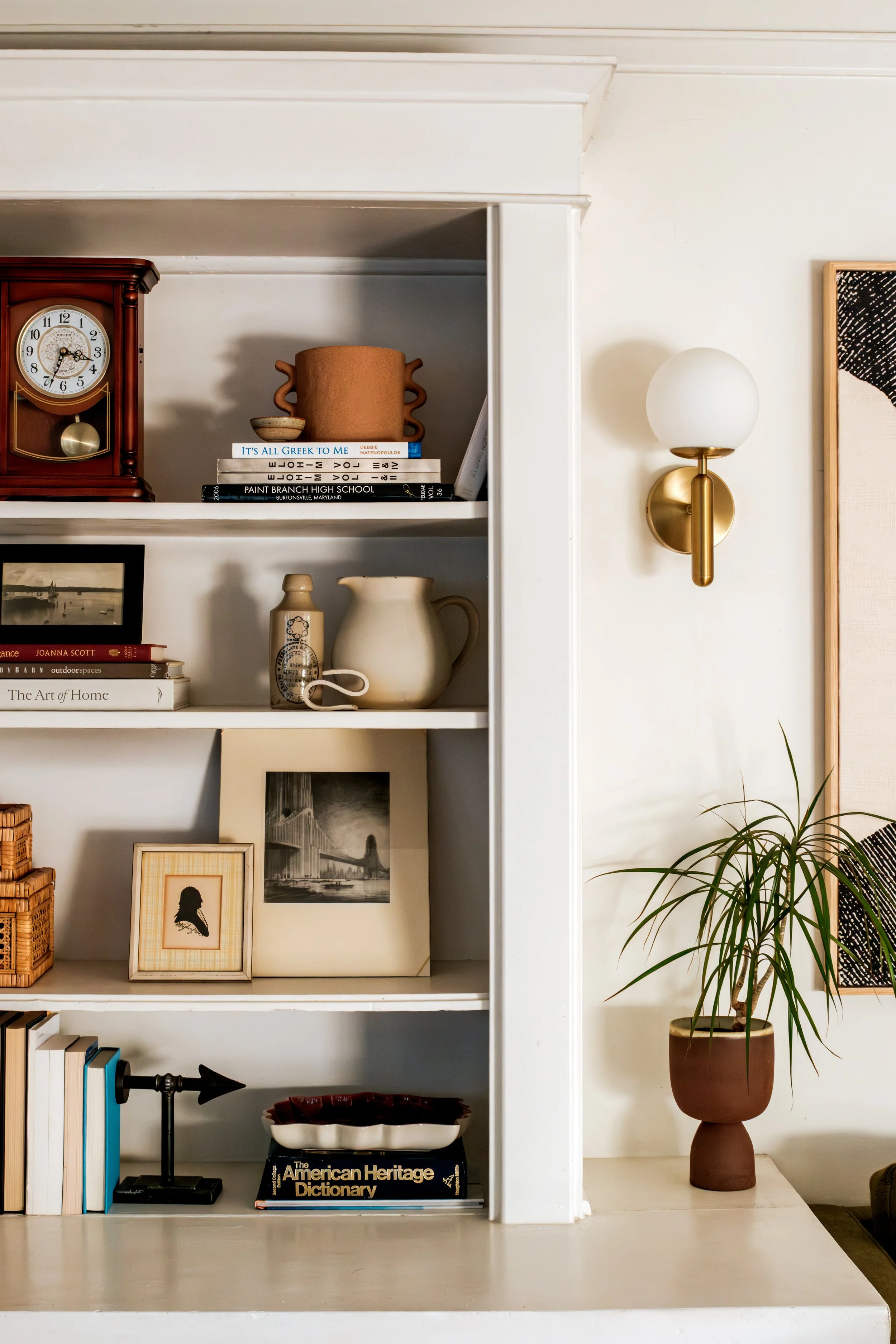 Part of a white bookshelf with decorative items, books, and photographs, a wall-mounted light fixture with a white globe shade, and indoor plants in a living room.