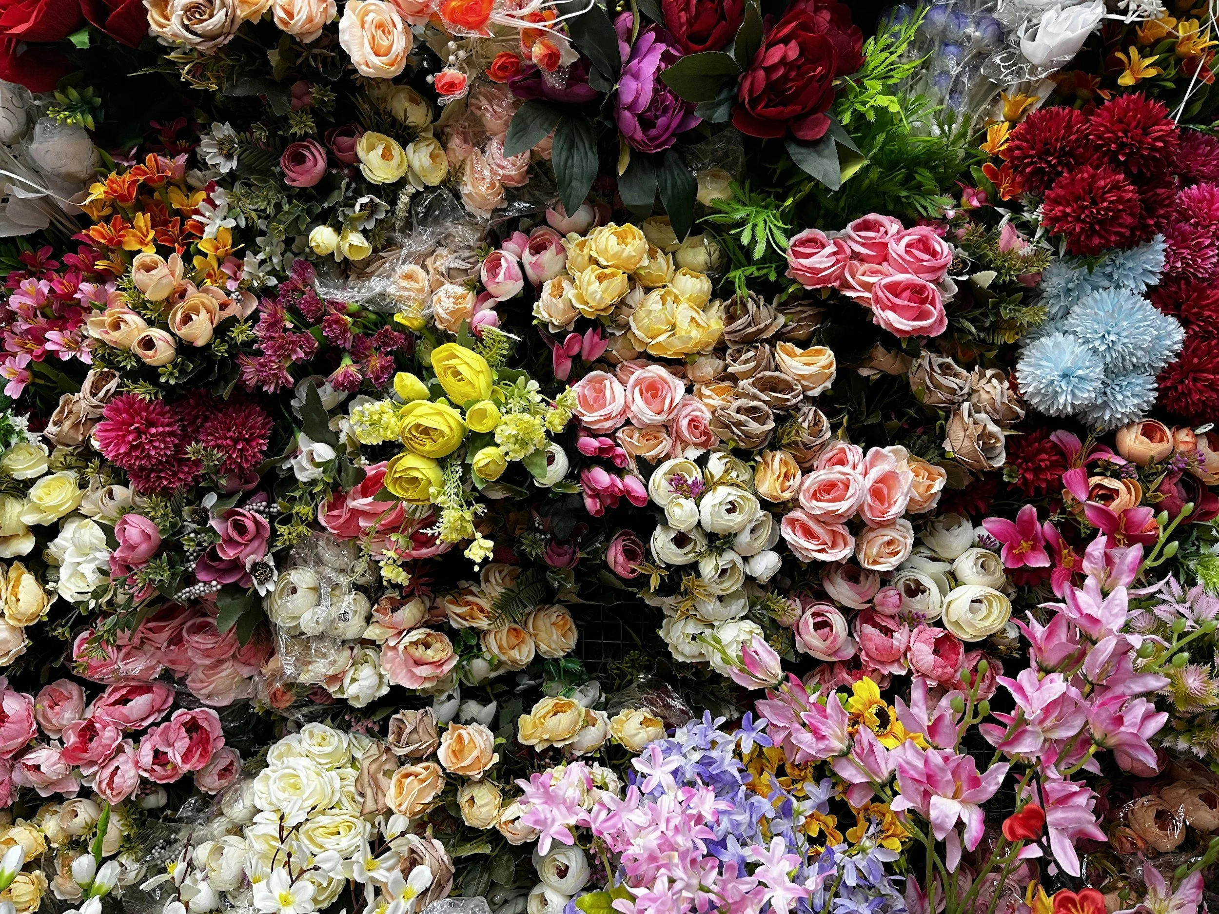 A colorful display of various artificial flowers, including roses, carnations, and lilies, arranged densely in a flower shop.