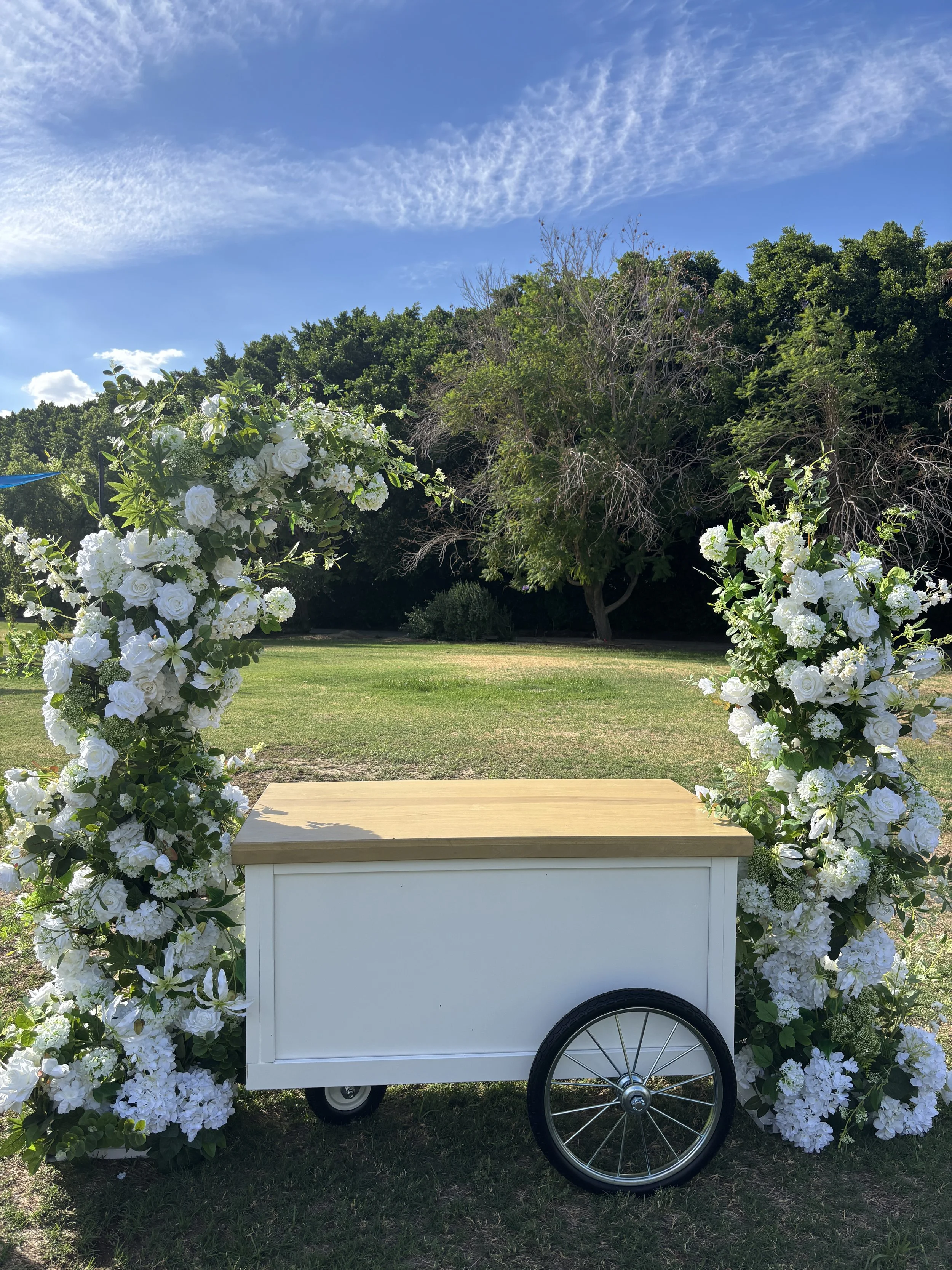 A small white cart with a wooden top, decorated with white flowers and greenery, set outdoors on grass with trees and a blue sky in the background.