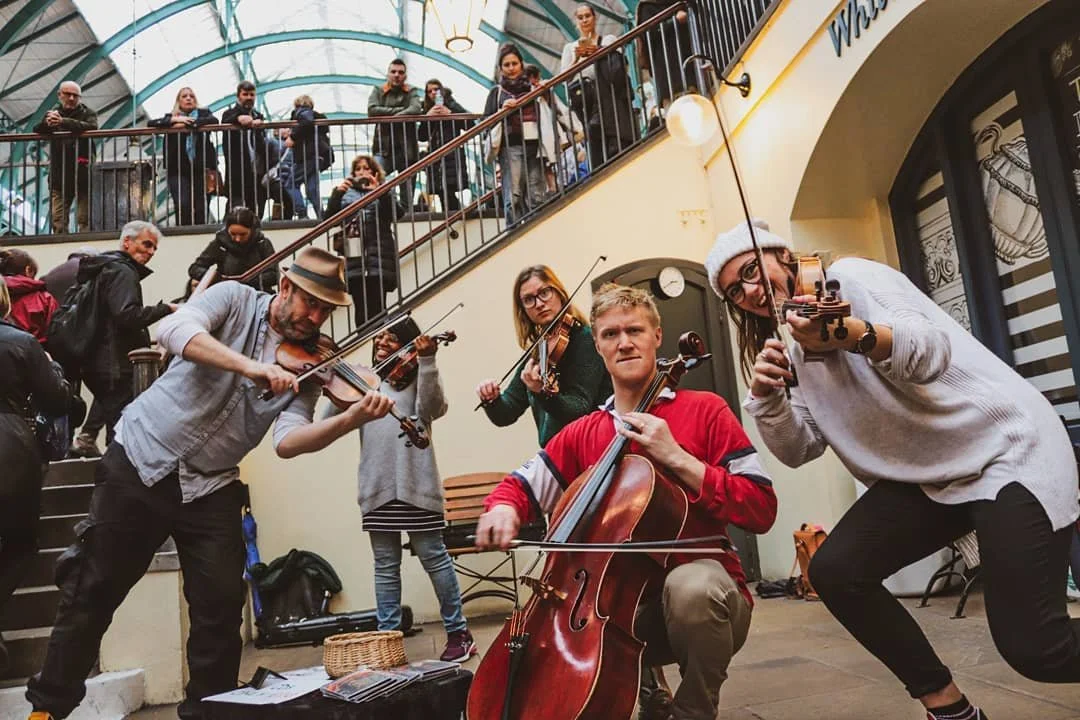 A group of five street musicians playing violins and a cello inside a shopping mall, with several onlookers watching from a balcony above. The ensemble is OM strings.