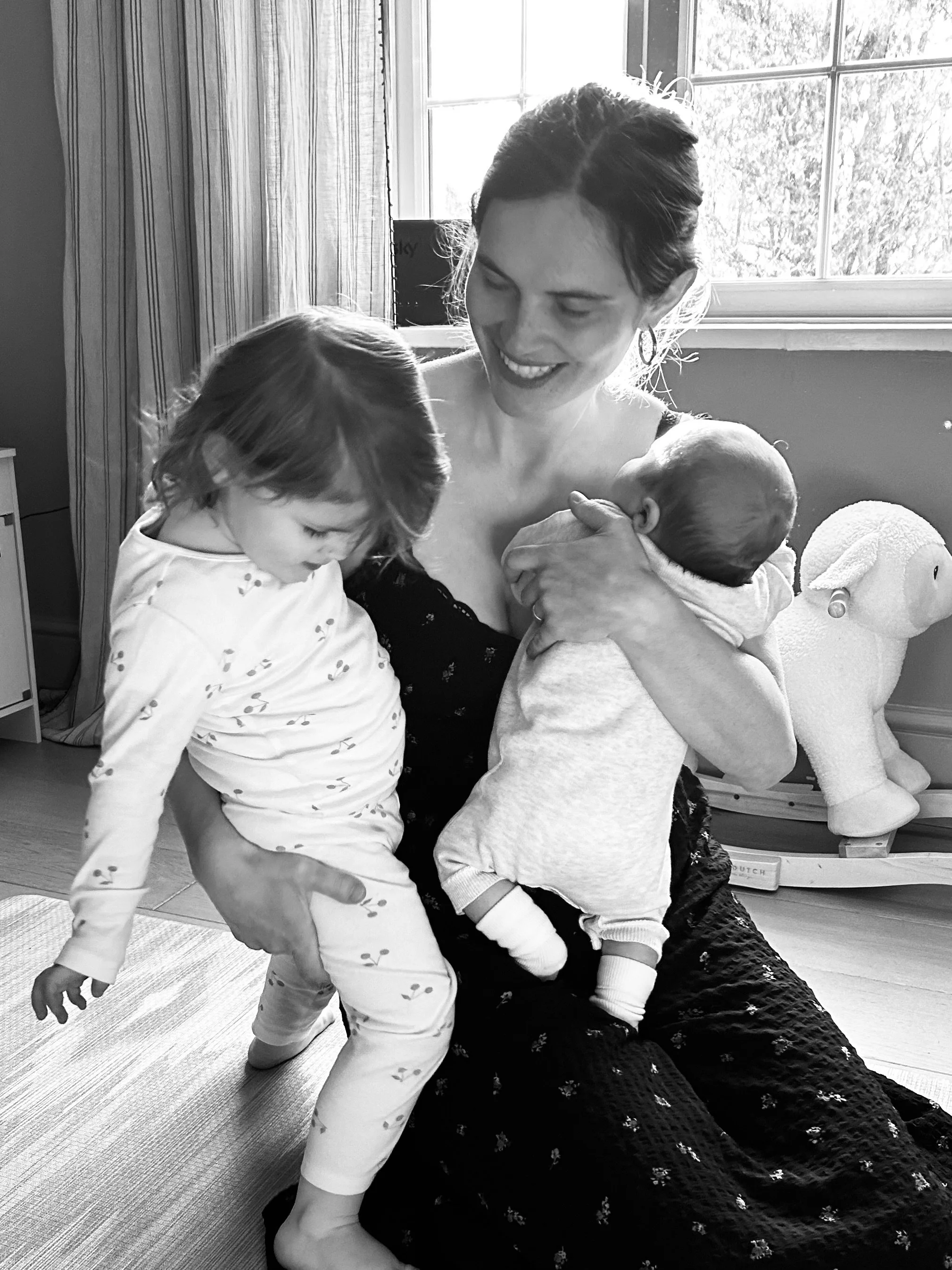 A woman smiling while holding two young children, one in each arm, in a cozy room with a window and wooden paneling. The children are leaning close to her, and the woman appears to be engaging with them affectionately.