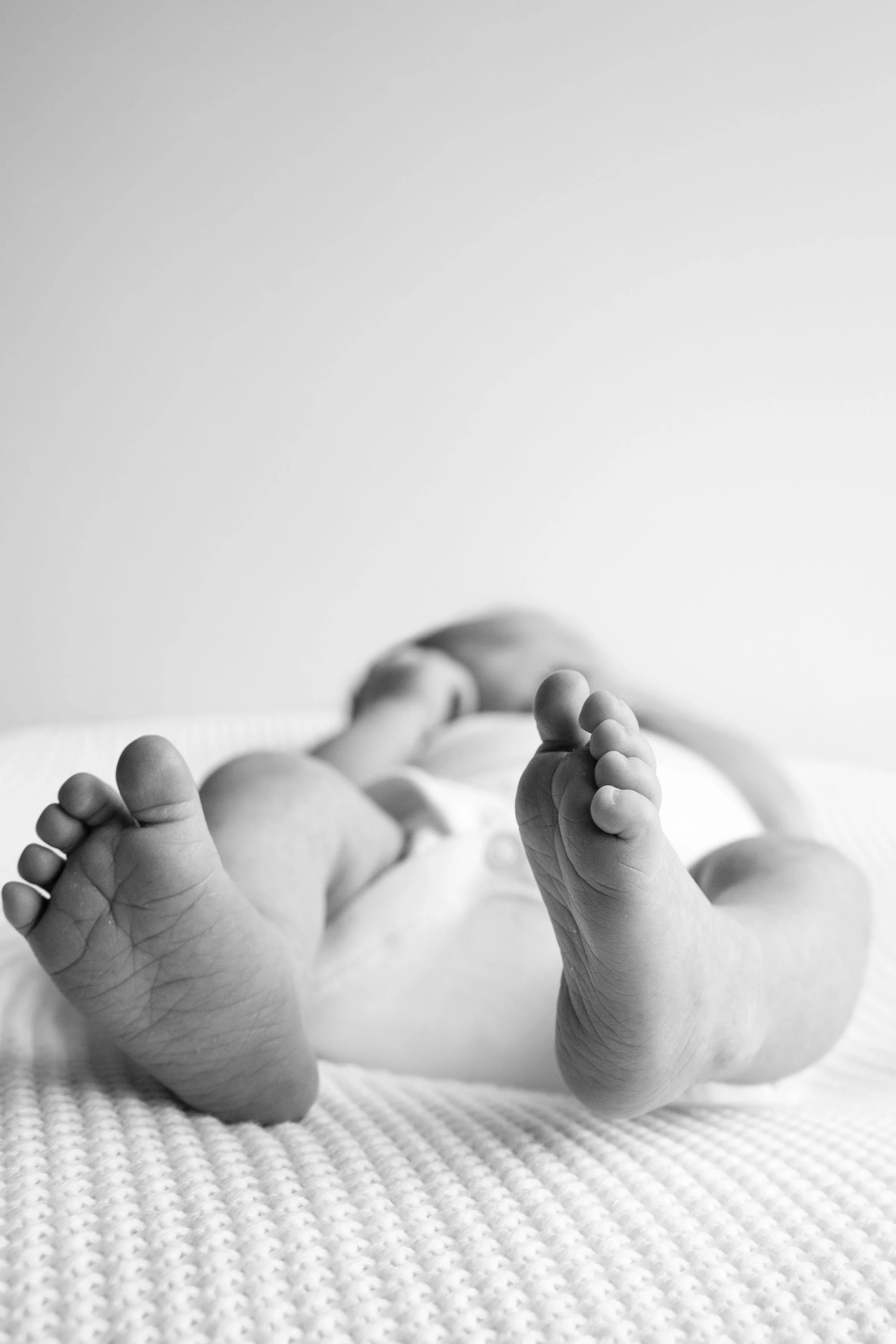 Black and white photograph of a baby's feet and hands, lying on a textured blanket.
