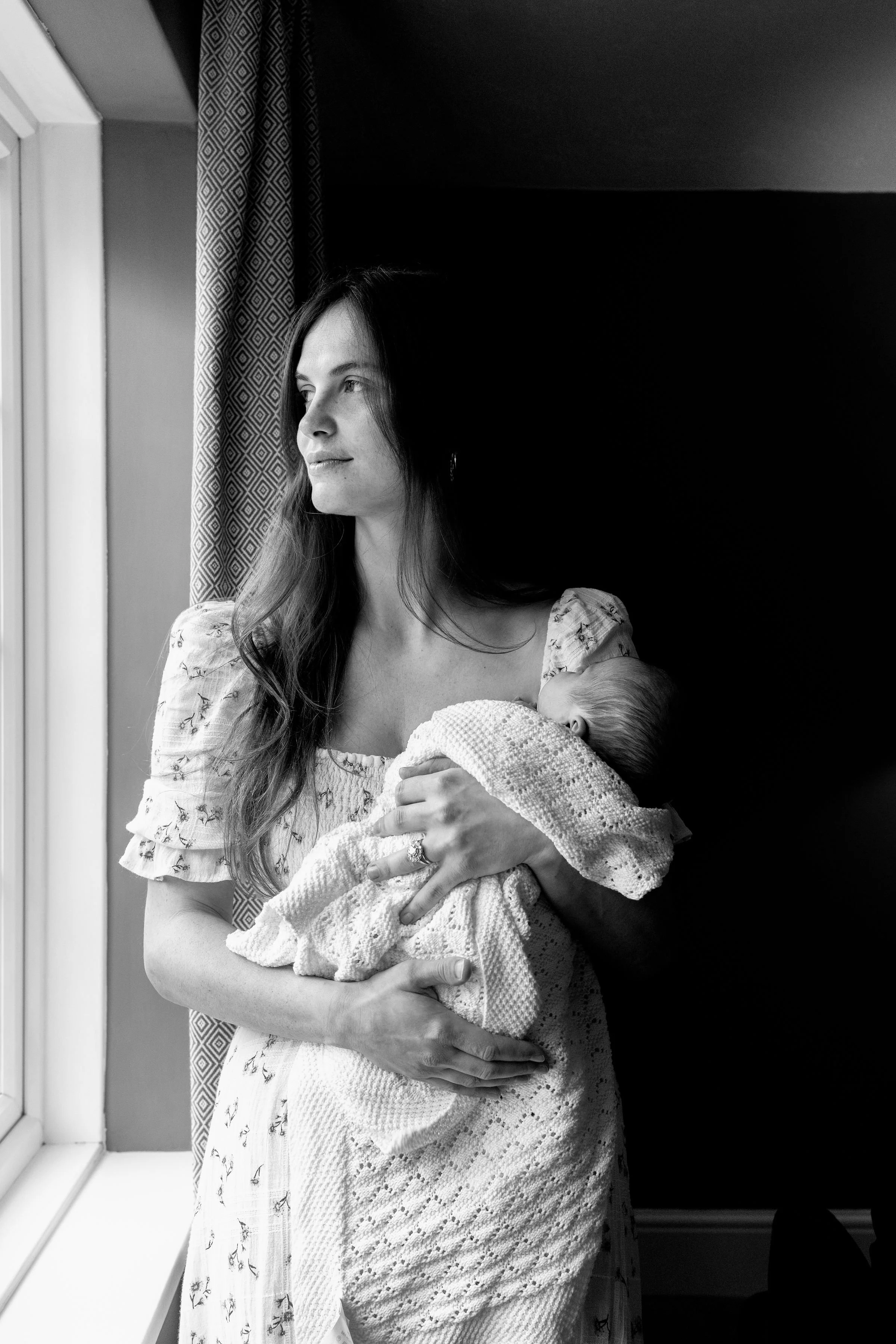 A woman with long hair holding a newborn baby near a window with curtains.