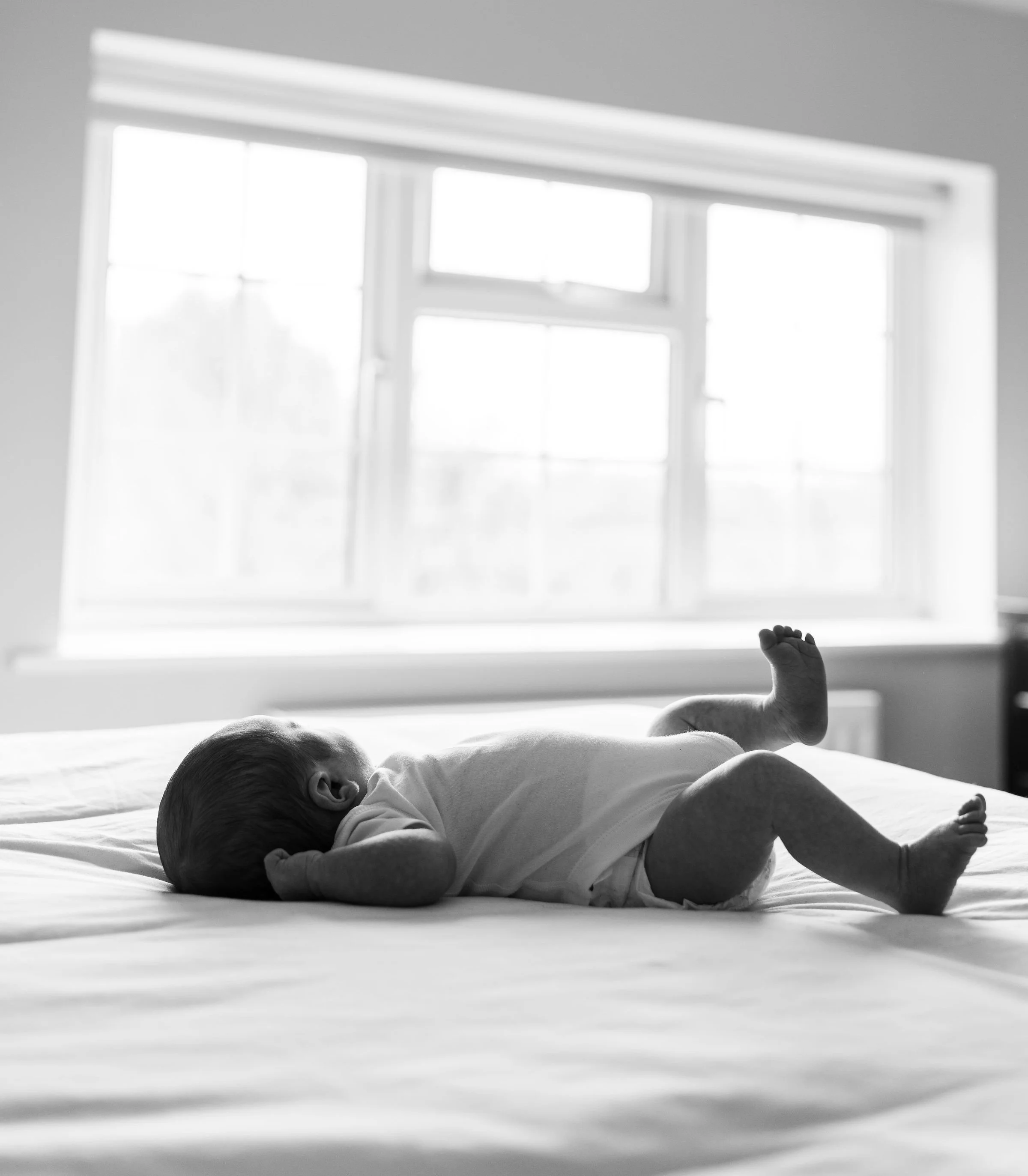 A baby lying on its stomach on a bed, reaching up with one arm, in a room with large window blinds letting in bright light.