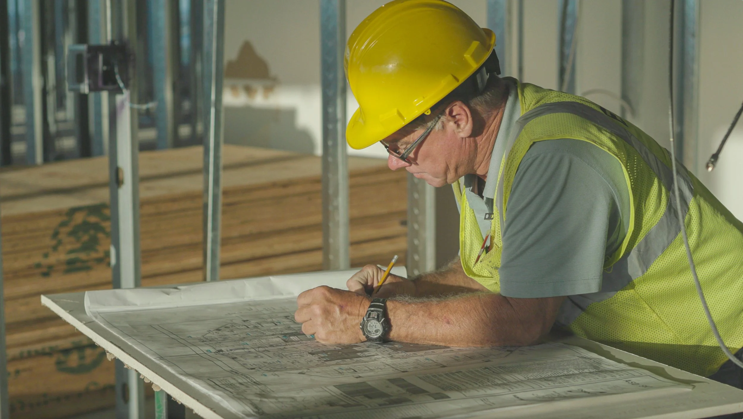 A construction worker wearing a yellow hard hat, safety glasses, and a high-visibility vest is leaning over a table with construction blueprints, writing or making notes with a pencil.