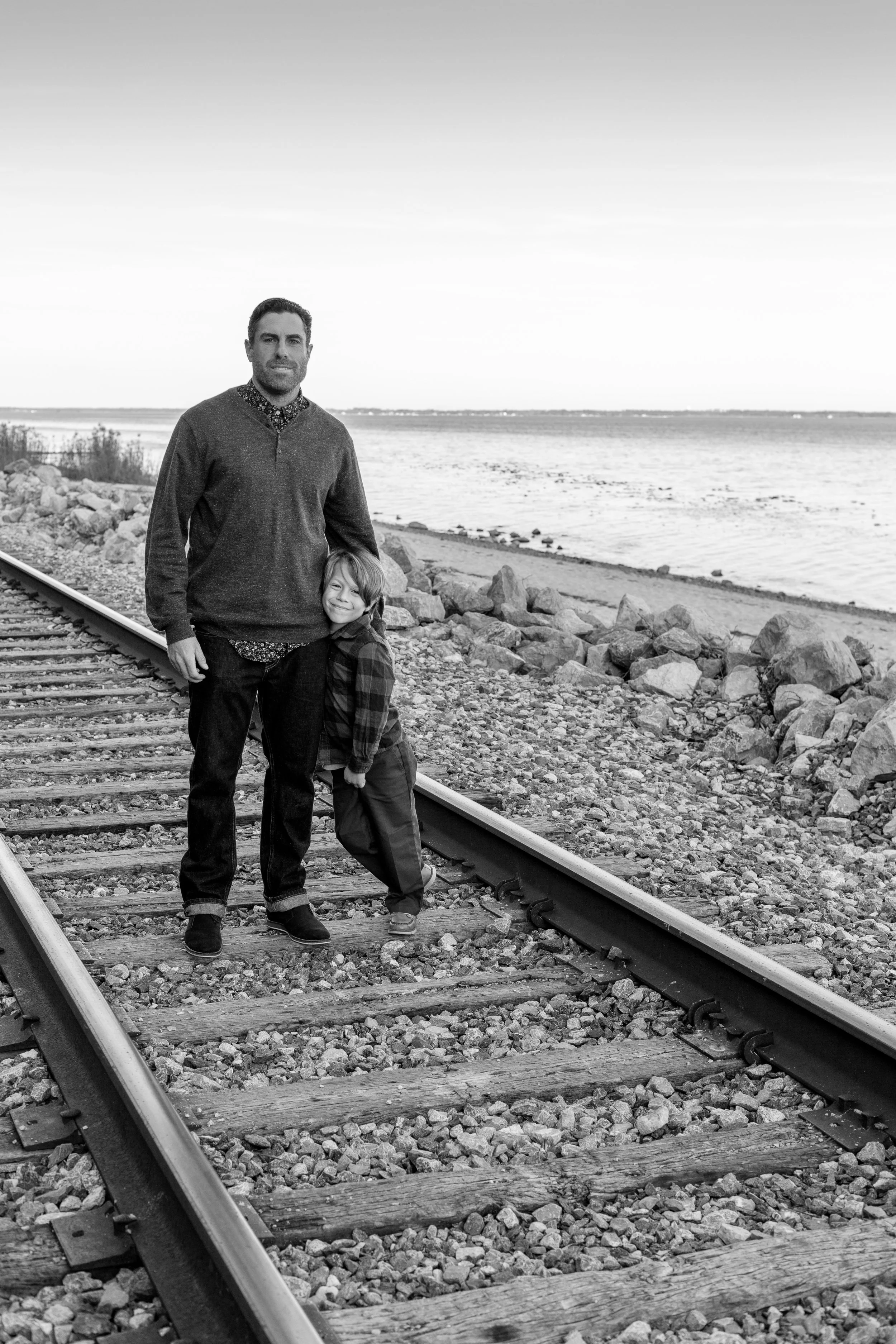 A man and a young boy standing on railroad tracks near a body of water, smiling at the camera, in a coastal or lakeside setting.