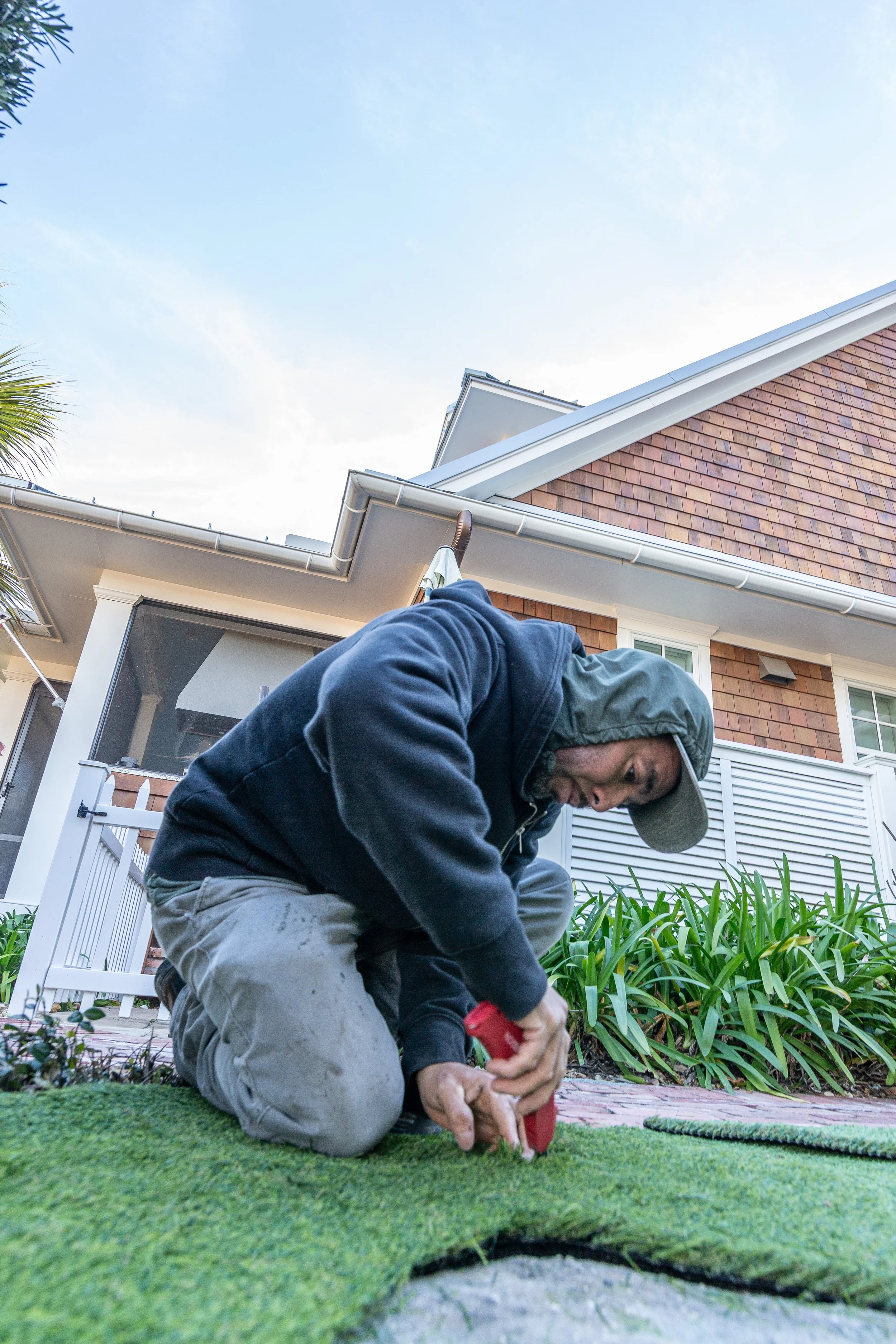 A man kneeling on the grass, using a red gardening tool, with plants and a house in the background.