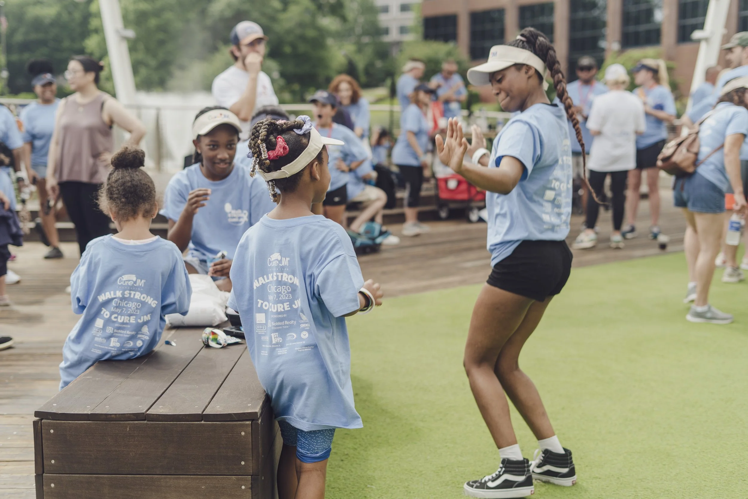 Group of children and adults gathered outdoors for a walkathon event, with some children wearing blue t-shirts that read 'Walk Strong Chicago May 7, 2023 To Cure JM,' engaging in conversations and activities.