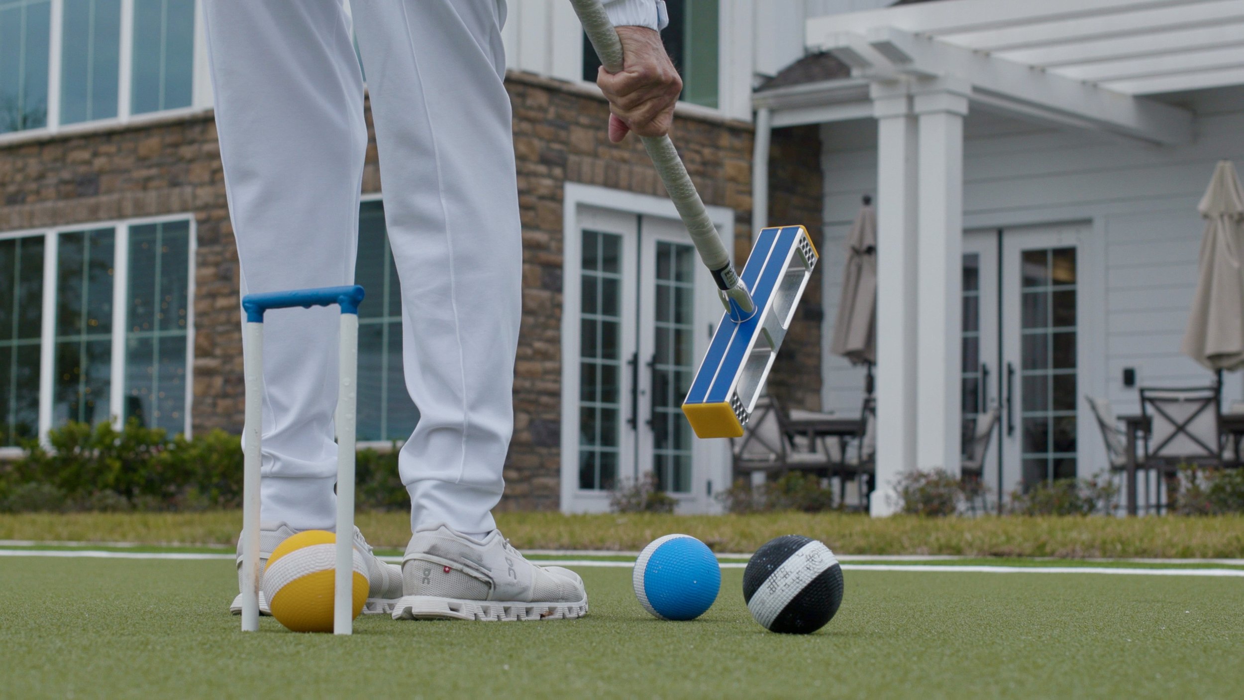 Person standing on artificial turf, holding a croquet mallet, with croquet balls and wickets around them, in front of a house with large windows and a porch.
