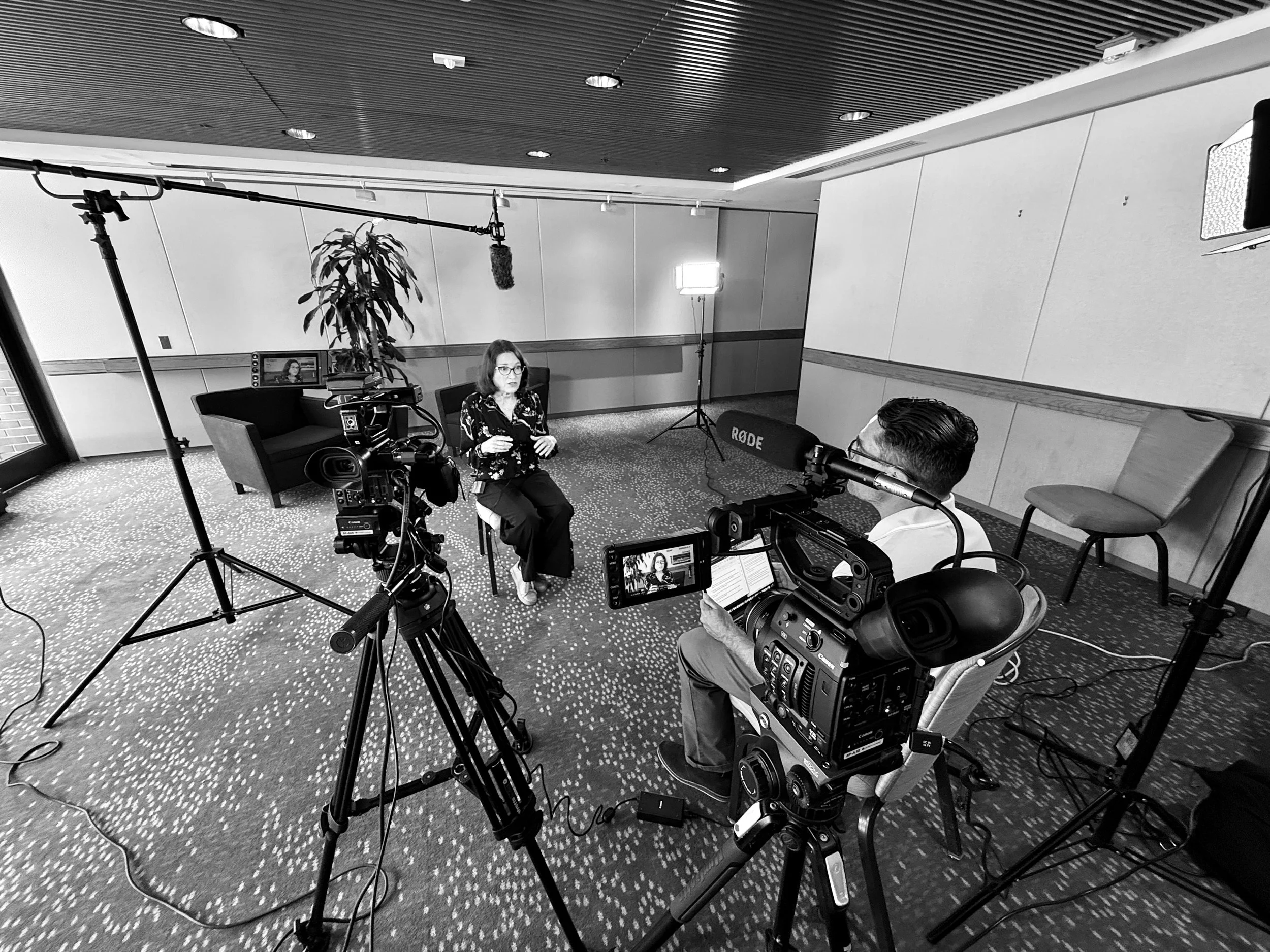 A woman wearing glasses being interviewed on camera, with a man operating recording equipment. Recording studio setting with lights, camera, and sound equipment.