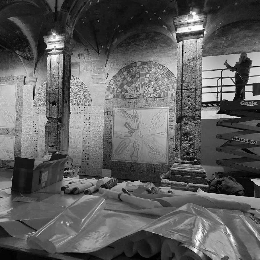 Interno di un'area espositiva con dipinti e opere in corso, scaffali con materiali di lavoro, una piattaforma di sollevamento con una persona che indossa un completo scuro