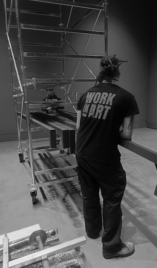 Una persona con dreadlocks indossa una maglietta con la scritta 'WORK IN ART' e lavora in un ambiente di officina o studio, con scaffali metallici e materiali da costruzione.