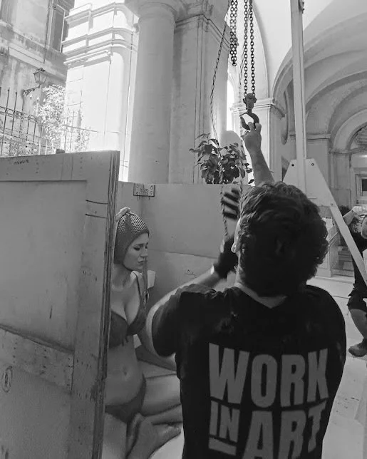 Artiista femminile che si prepara davanti a un set di produzione, con un membro della troupe che regge una luce o un dispositivo di illuminazione, all'interno di un edificio storico o governativo.