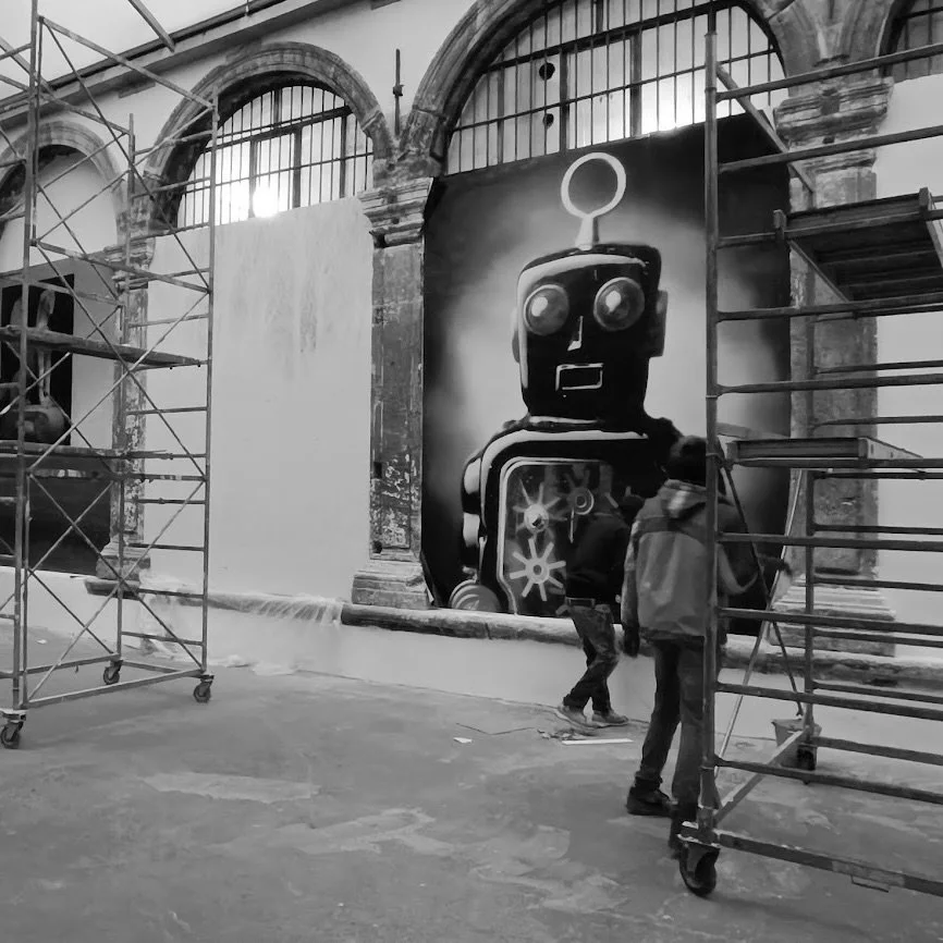 Un mur con un grande murale di un robot stilizzato, in un ambiente con scaffalature e strutture di supporto allestite in un edificio con archi in pietra.