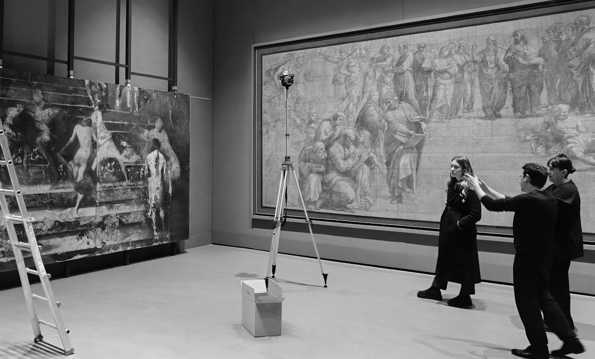 Tre persone in un museo davanti a due grandi opere d'arte, una moderna raffigurante bambini che giocano con l'acqua e un'altra classica con figure umane. Disegnatrici o guide stanno spiegando le opere.