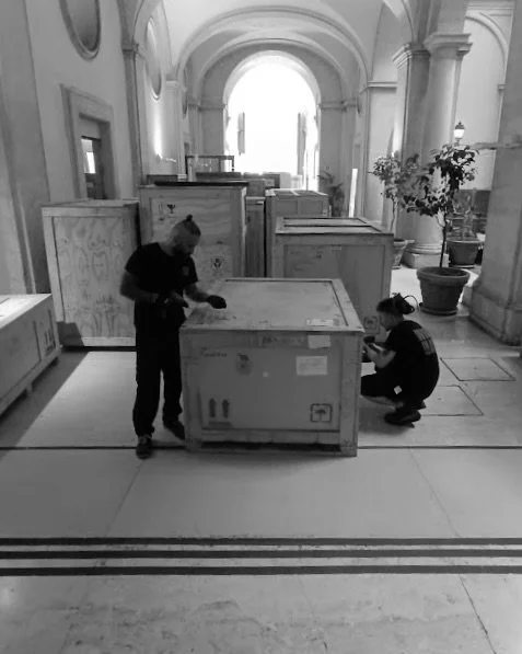 Due persone che ispezionano grandi casse in un'architettura classica con archi e colonne, all'interno di un edificio storico.