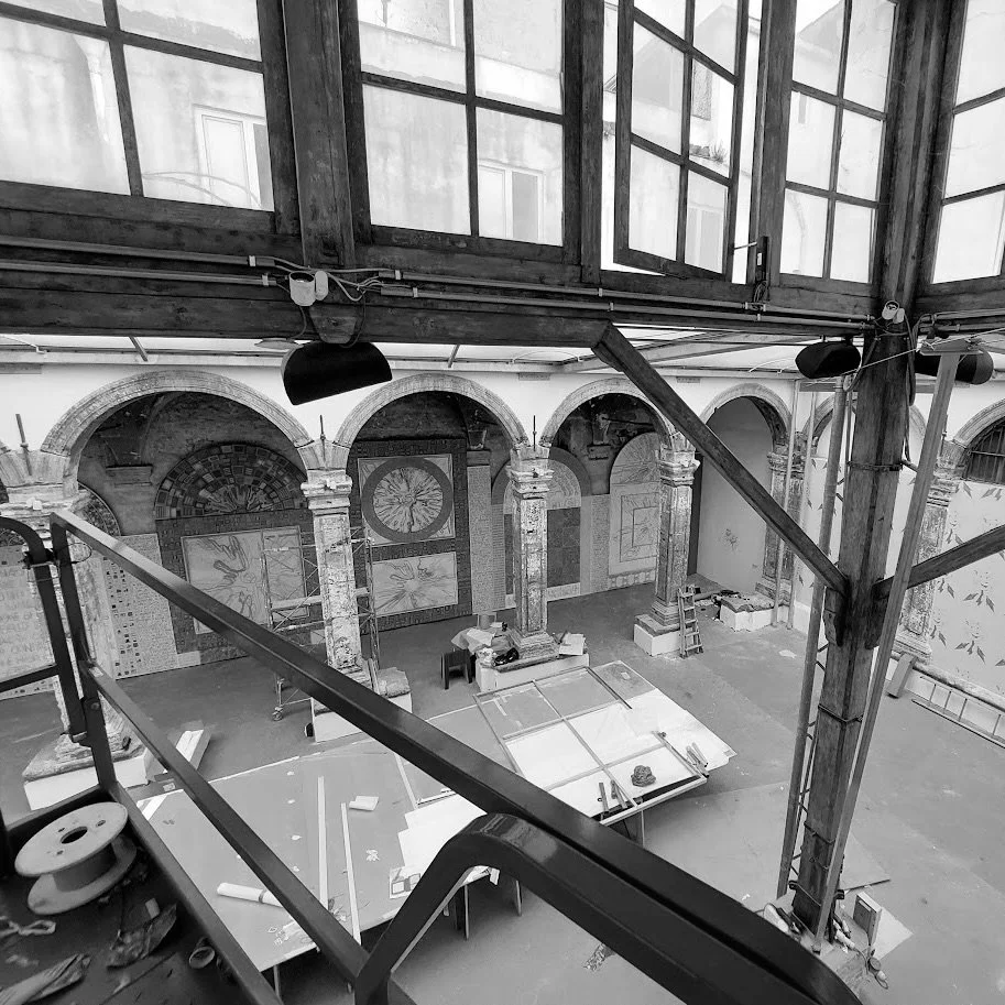 Interno di un edificio storico in ristrutturazione, con scaffalature, scala e pareti decorate con affreschi o mosaici.