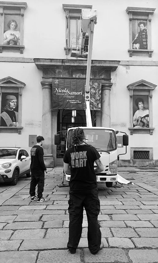 Personas che si trovano davanti a un camion con carro elevatore sollevato, di fronte a un edificio storico con dipinti alle finestre. Una persona indossa una maglietta con scritto "WORK IN ART".