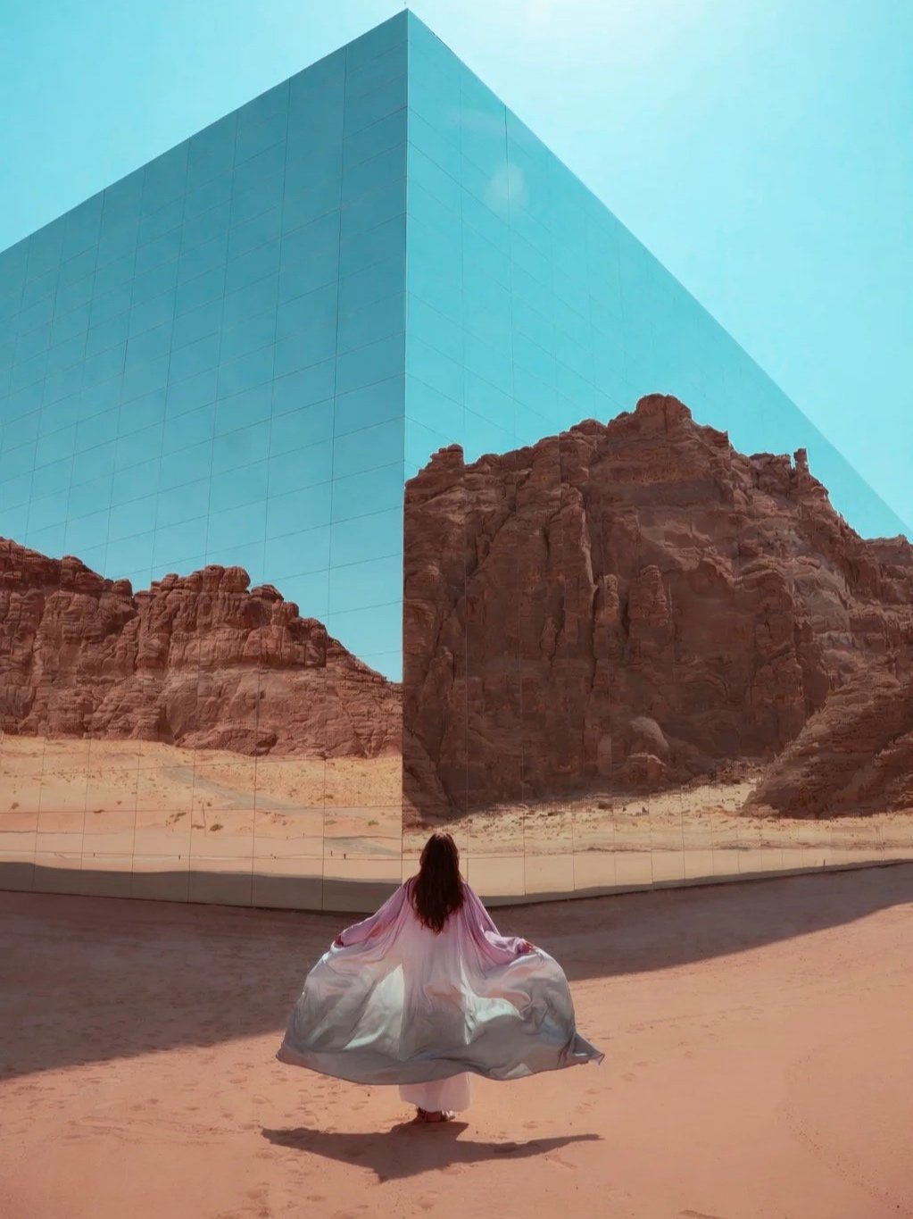 A woman in a flowing pink and gray dress stands in front of the world's largest mirrored building, Maraya, in AlUla. The screen creates an illusion of an extended desert scene behind her.