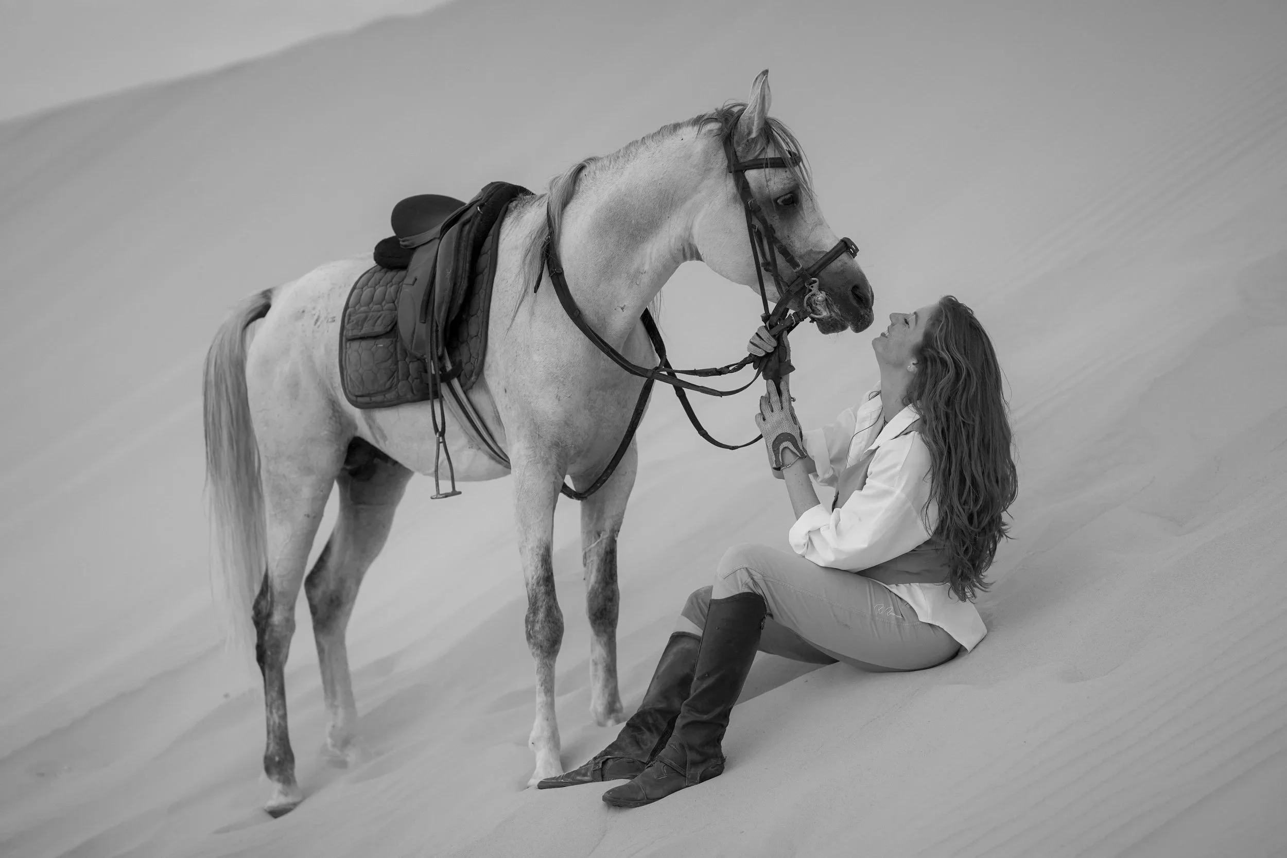 A woman with long hair sitting on the sand in front of a horse, smiling while holding the horse's bridle, in a desert environment.
