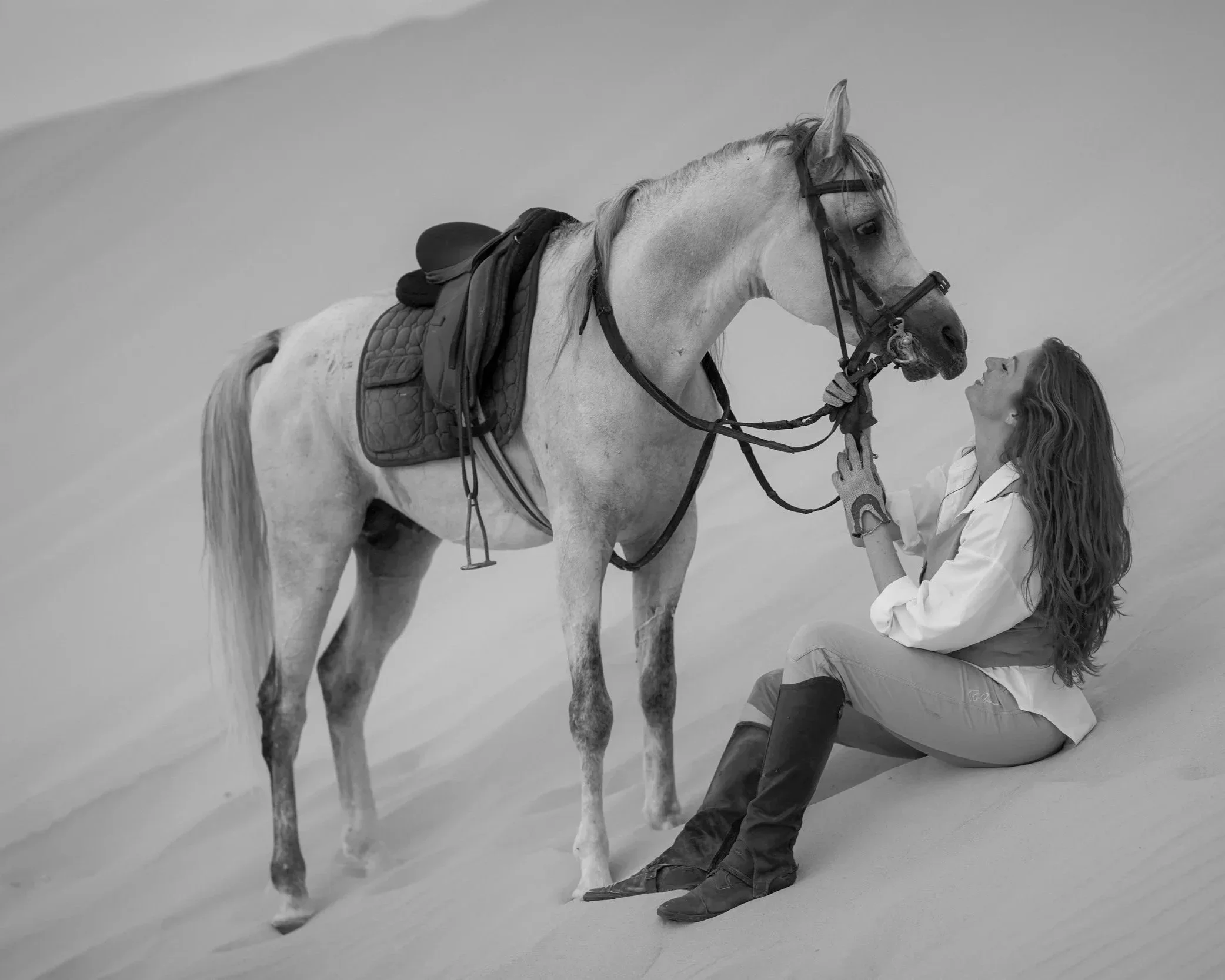 A woman sits on the sand in front of a horse in a desert landscape, sharing a joyful moment and looking at each other.