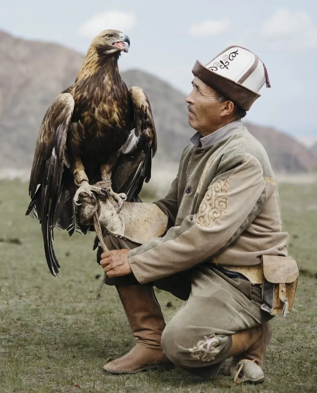 A Kyrg man in traditional Central Asian clothing and a tall hat kneeling on the grass, holding a large bird of prey, a golden eagle, on his gloved arm, with mountains in the background.