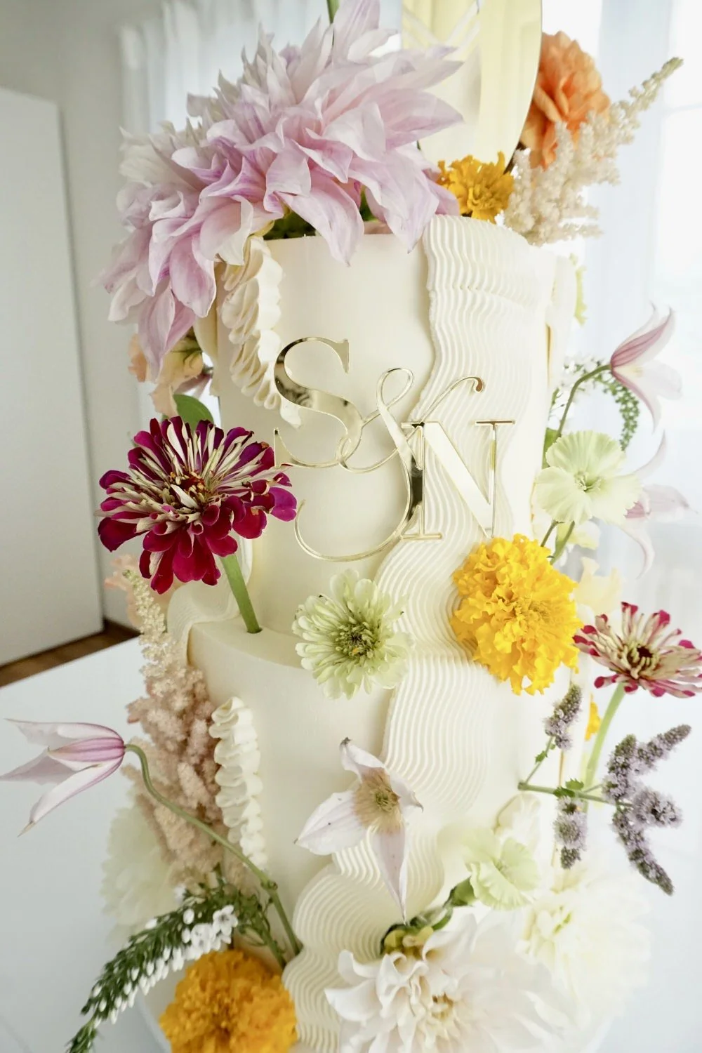 A multi-tiered wedding cake decorated with colorful flowers including pink, yellow, white, and maroon blossoms, and featured with gold letters S & N on the front.