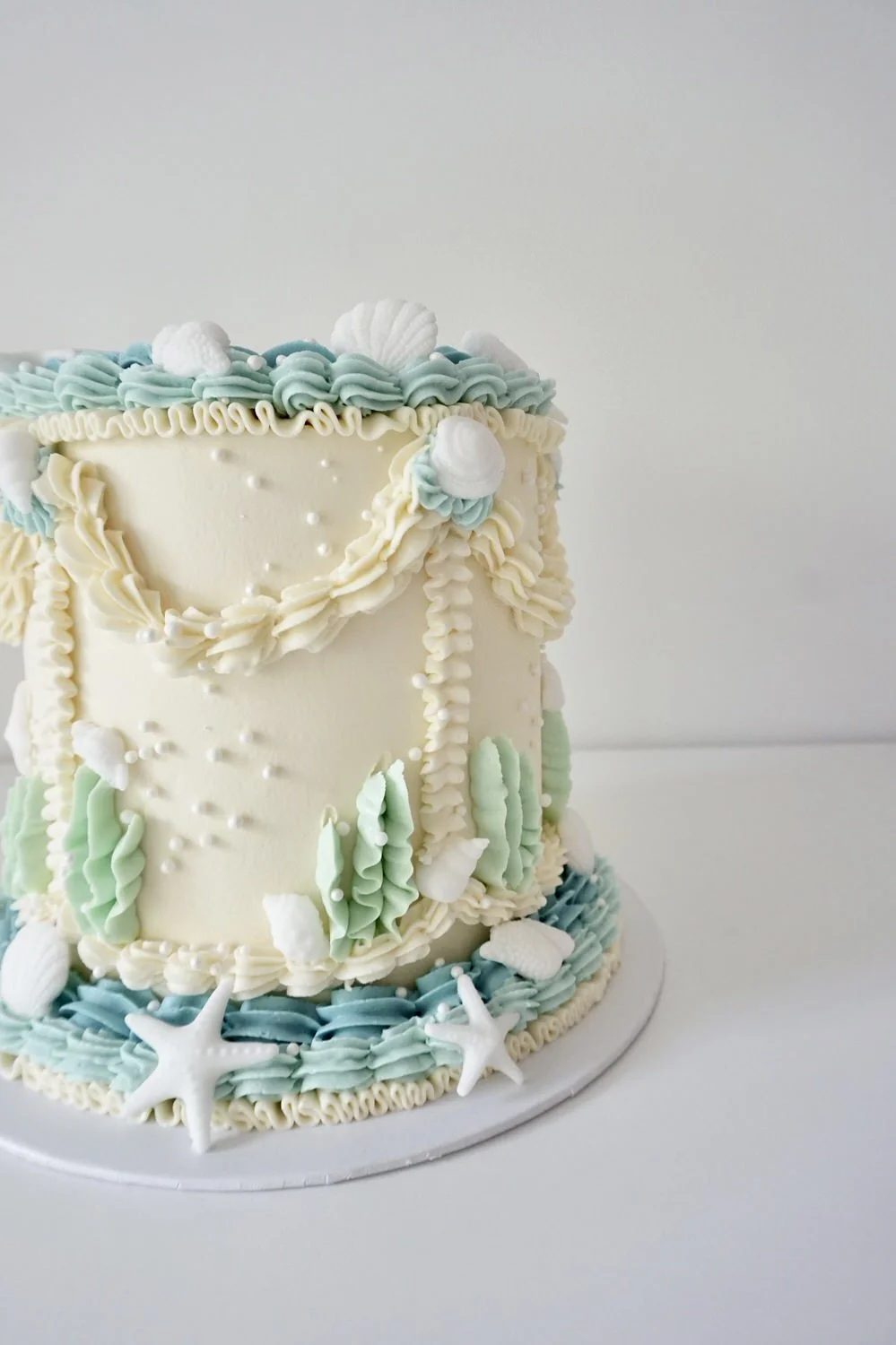 A seaside themed decorated cake with white, blue, and green piped frosting, shaped like seashells, starfish, and seaweed.