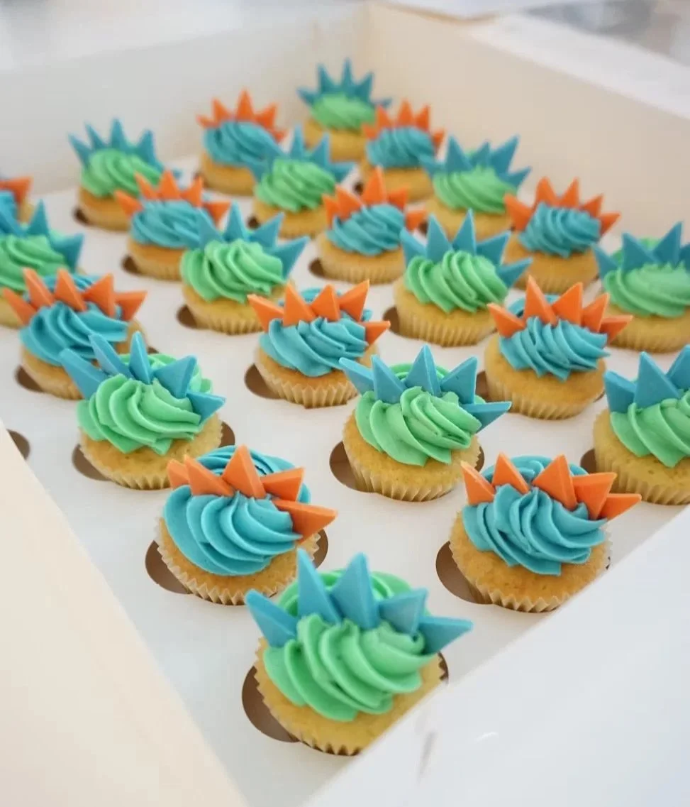 A box of cupcakes decorated with green, blue, and orange frosting and triangular paper decorations.