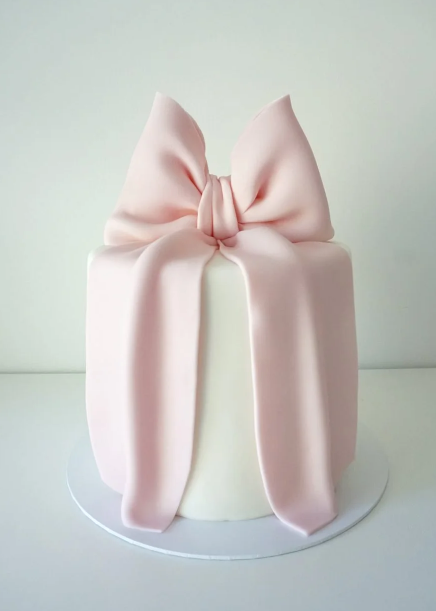 A cake decorated with a large pink fondant bow on top, with long tails cascading down the sides, set against a plain background.