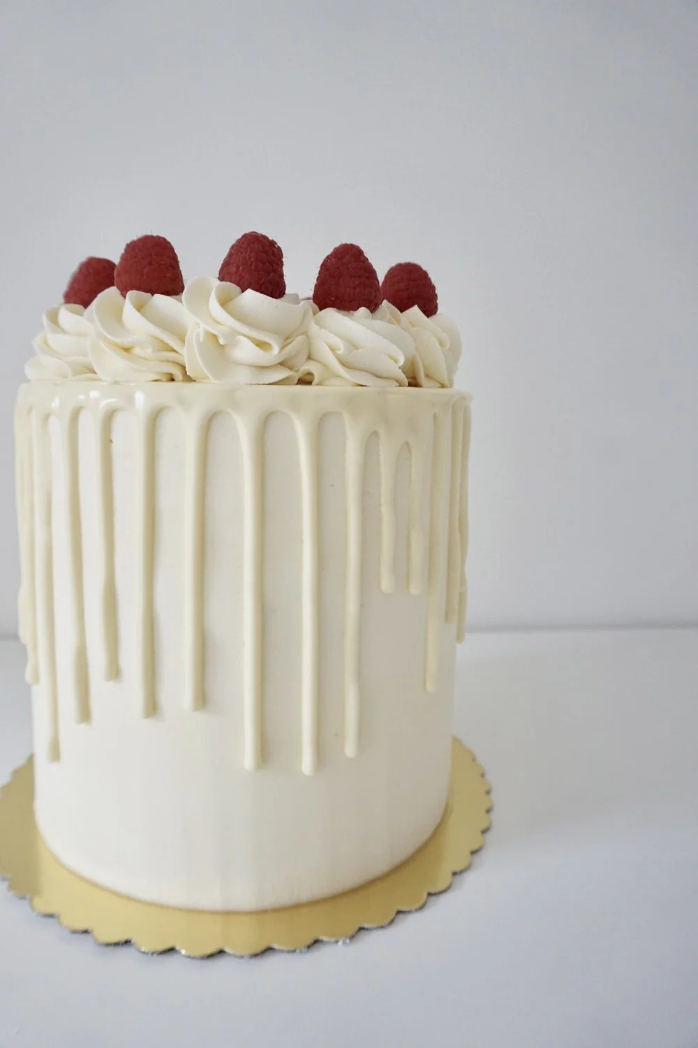 Elegant cake with white walls, white chocolate drip decoration, topped with whipped cream swirls and fresh raspberries.